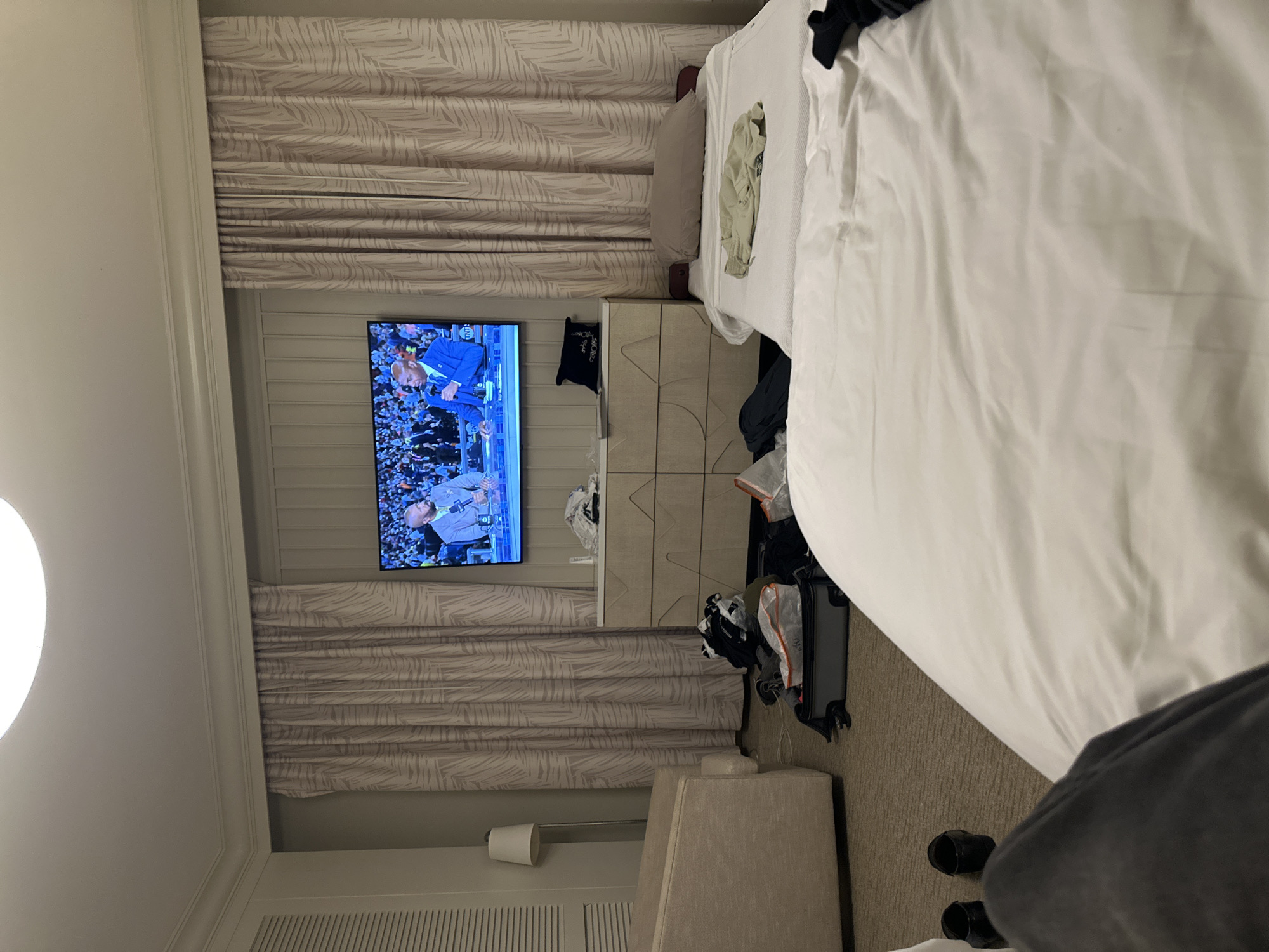 A bedroom in Aventura, United States of America, contains a made bed with white linens, positioned in the right side of the frame, covered with scattered clothing. A television mounted on the wall is displaying a sporting event in front of a closed curtain. To the left of the TV, a dresser is loaded with assorted items, including garments. Adjacent to the bed, on the right, is a suitcase lying open, along with various other belongings scattered on the floor. In the lower right corner, a portion of an individual’s legs and shoes are visible. The room is illuminated by artificial lighting.
