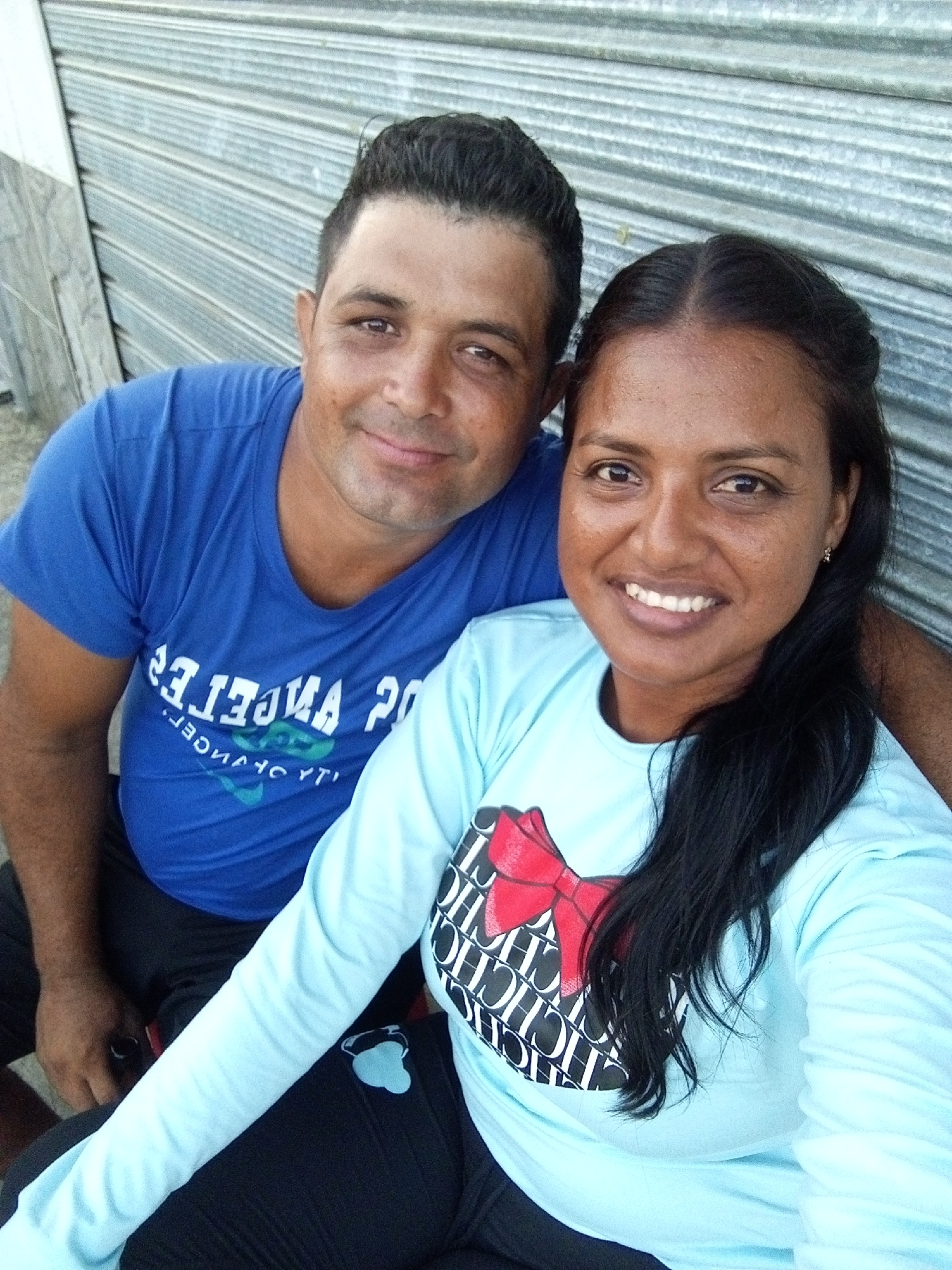 A close-up photograph shows a smiling man and woman posing for a selfie. The man is wearing a blue t-shirt with the text "Los Angeles 20" printed on it. The woman is wearing a light blue long-sleeved shirt with a red and black bow and text design. They are sitting close together, with the man having his arm around the woman. Behind them is a corrugated metal wall. The location is Unknown, Unknown.