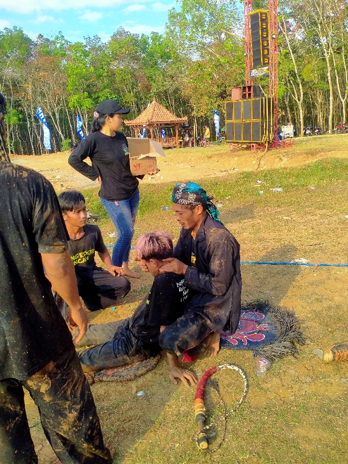 The image depicts an outdoor scene in Kuto, Indonesia, featuring individuals and structures suggestive of a cultural event or performance. In the foreground-center, a male individual with pink-dyed hair, wearing mud-splattered black attire, leans forward on the ground. Directly behind him, an older male, also in heavily soiled dark clothing and a blue and white patterned headwrap, sits on a dark, patterned mat. His hands are placed on the shoulders and upper back of the younger man, appearing to offer support. To their left, a male in a black t-shirt and dark pants kneels on the ground, observing the central interaction. Standing slightly to the left and behind the kneeling man is a female wearing a black long-sleeved shirt, blue distressed jeans, and a black baseball cap; she holds an open cardboard box and is bare-footed. A partially visible male figure, also covered in mud, stands in the far left foreground. On the ground in the lower right foreground, a whip with a reddish-brown, yellow, and black handle and a braided white thong is visible alongside a small clear plastic bottle. The ground is primarily dry, light brown soil with scattered patches of green grass. In the mid-ground, a traditional Indonesian-style gazebo with an orange tiled roof is visible to the left, and a tall, multi-tiered sound system array on a yellow metal scaffold stands to the right. Blue and white flags are visible in the background, which is dominated by numerous trees with light-colored trunks under a bright blue sky.