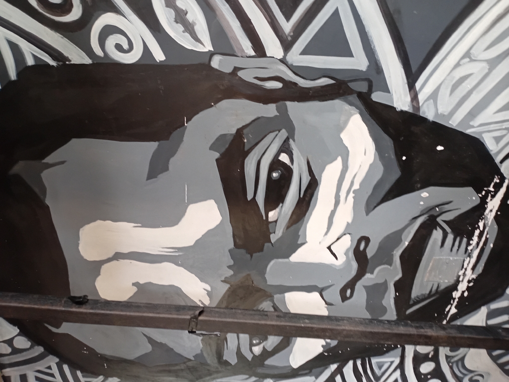 This is a grayscale mural depicting a close-up of a man's face, rendered in a bold, graphic style. The mural is located on a wall in the city of Howayi, Nigeria. The man's eyes are wide open, with prominent shadows defining his facial features. A vertical dark metal pole runs down the left side of the image, obscuring a portion of the mural. The background of the mural features abstract patterns, including swirls and geometric shapes in shades of black, white, and gray, creating a dynamic contrast with the central portrait. The paint application is somewhat rough, with visible brushstrokes and splatters, suggesting an outdoor or street art context.