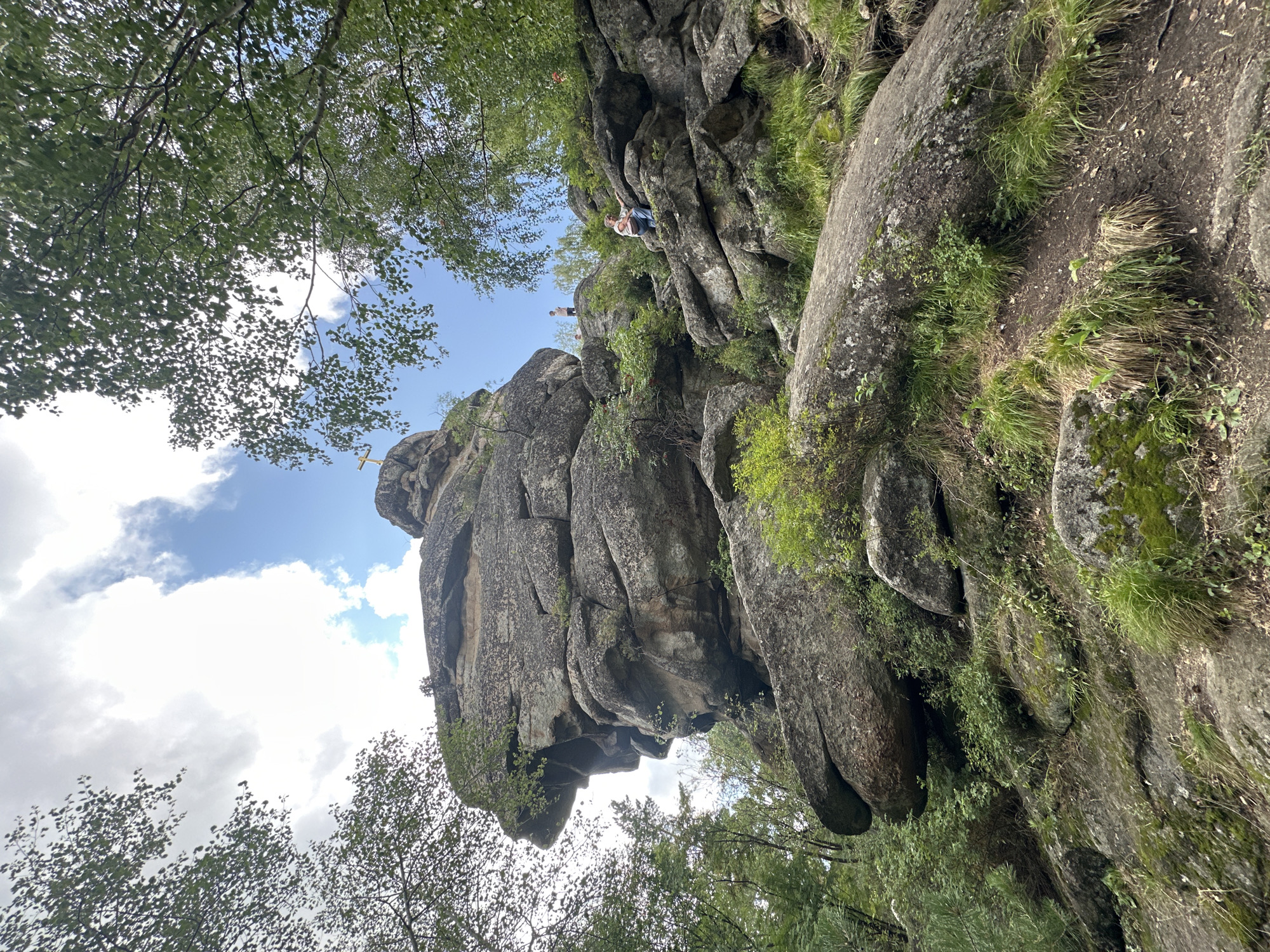 The image captures a natural scene in the city of Belokurikha, Russia. A prominent rock formation, resembling a human profile, dominates the center of the frame. The rock is massive and appears to be made of rough, layered stone. Lush green vegetation, including trees with dense foliage and smaller plants, surrounds the rock formation. In the background, the sky is visible with scattered clouds and patches of blue.
On the right side of the rock formation, two individuals are present. One person, appearing to be a woman wearing a pink top and blue jeans, is seated on the rocks. Another, taller individual, possibly a man, is standing further up the rocky slope. A small golden cross is visible atop a distant peak, suggesting a religious or memorial significance to the location. The terrain is rocky and uneven, indicative of a mountainous or hilly environment. The overall impression is one of a scenic, outdoor landscape with natural geological features and human presence.