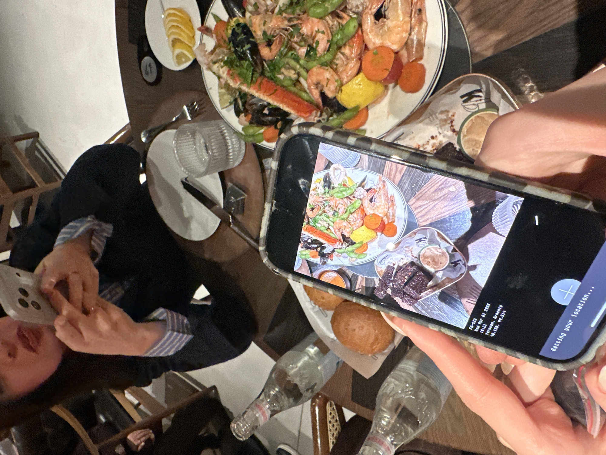 Here is a detailed description of the image:
The image captures a dining table setting, where a person is holding a smartphone, displaying a photo of the food laid out on the table. On the table itself, there is a large platter with seafood including shrimp, mussels, crab legs, asparagus, and sliced carrots and lemon wedges. There's also a smaller dish with lemon slices, a plate with what appears to be a side dish, and a clear glass.
Adjacent to the smartphone in the foreground, there are rolls and a dish with what looks like a dip and some protein (possibly meat). There are also two partially visible glass bottles.
A person is partially visible in the background, appearing to use another smartphone.
The smartphone in the foreground is displaying an image of all the food on the table. Text visible on the phone's screen indicates a date of "Wed Apr 01 2026, 16:21," and the location "Yerevan, Armenia" with coordinates "48.198, 44.514." The phone also reads "Getting your location..."
