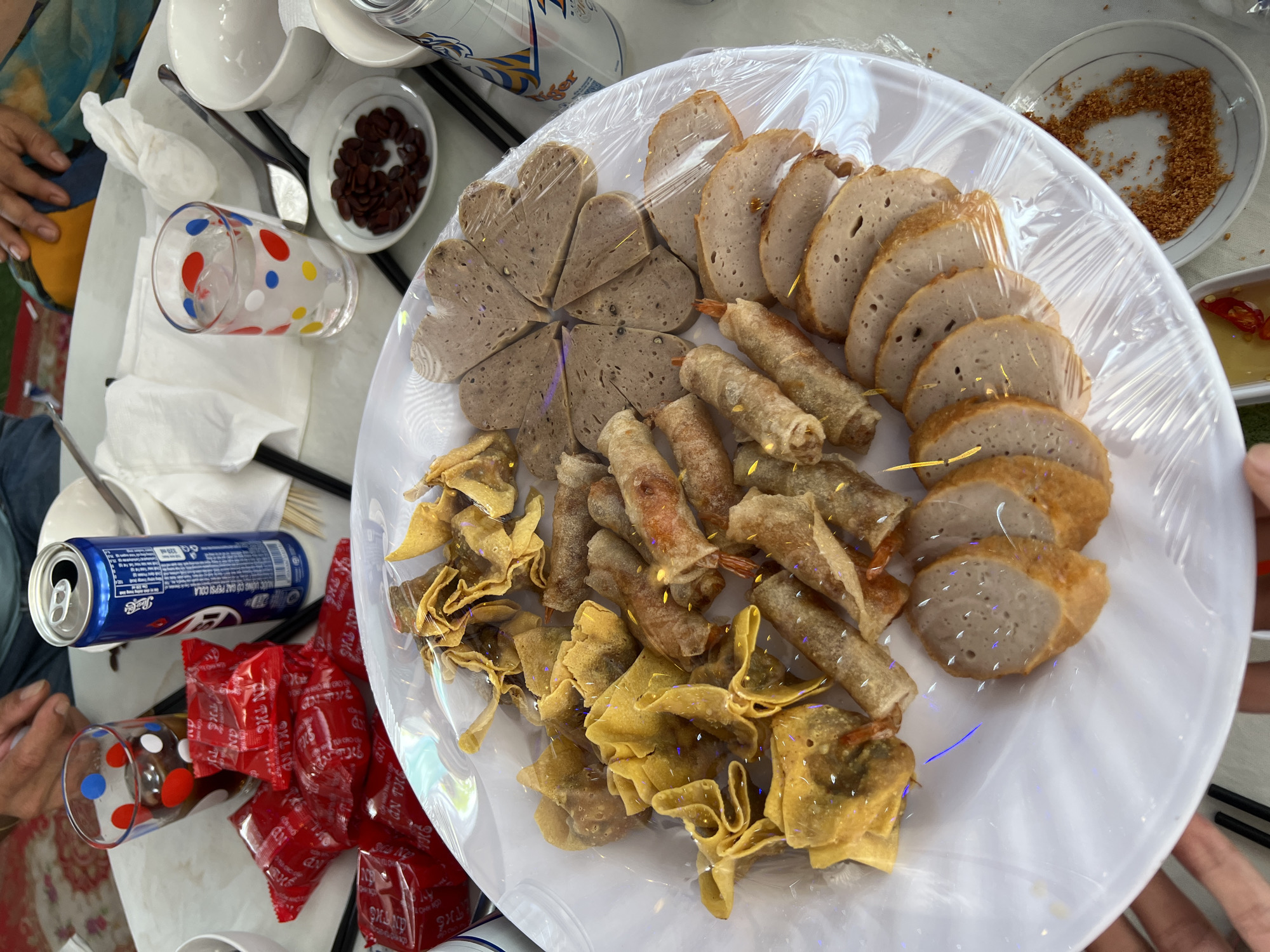 A high-angle shot captures a casual meal spread on a light-colored table, likely in Bình An, Vietnam. The central focus is a large white platter, covered with transparent plastic wrap, laden with various Vietnamese dishes. These include heart-shaped and round slices of a light brown, sausage-like item (possibly chả lụa or giò lụa), fried spring rolls, and crispy fried wonton-like snacks.
Around the platter, several hands are visible, suggesting people are gathering for the meal. Beverages include an open blue Pepsi-Cola can and a silver Tiger Beer can, along with a clear glass with colorful polka dots holding a dark drink. Tableware consists of white bowls, small plates (one containing dark reddish-brown seeds, another with what appears to be toasted coconut or peanut flakes), chopsticks, and white napkins. A small dish holds dipping sauce with sliced red chilies.
Next to the Pepsi can are several small red packets, likely containing condiments or snacks, with Vietnamese text such as "CÒN NHỚ THU CHO CẤP" and "AN THO". The Pepsi can itself has "NƯỚC UỐNG CÓ GAZ PEPSI COLA" printed on it. The bright lighting suggests it is daytime. The entire scene depicts a convivial gathering enjoying a selection of local Vietnamese food.