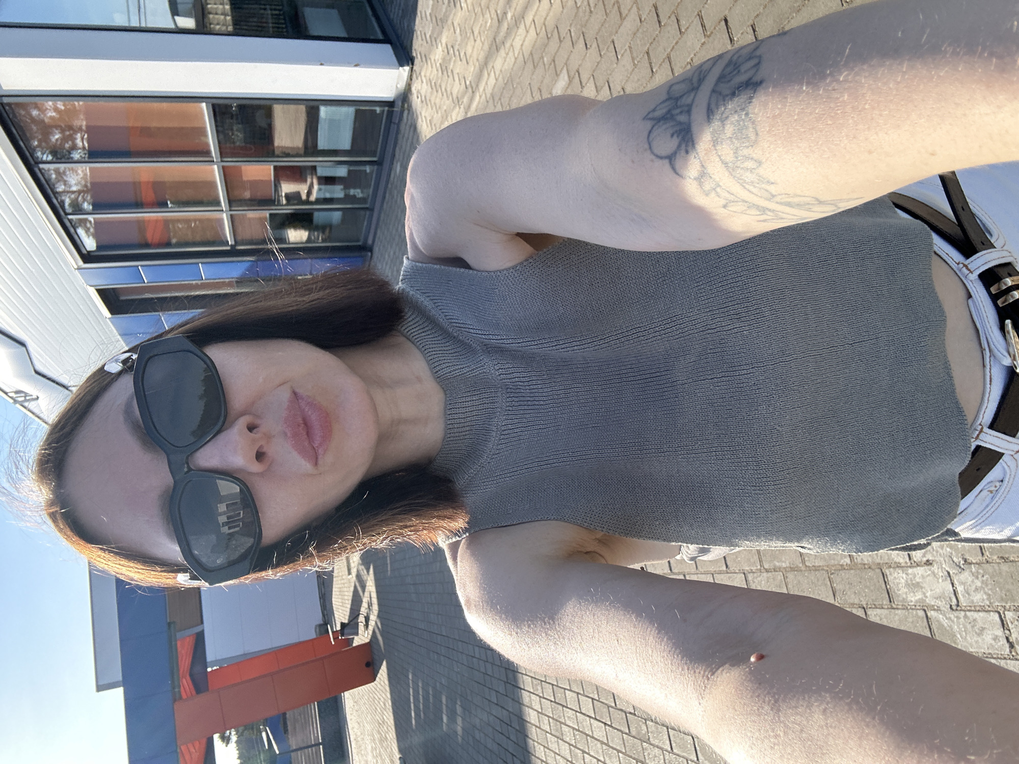 A selfie taken by a light-skinned woman with shoulder-length brown hair, wearing black sunglasses and a gray knit top with a white belt and pants. The woman's right arm, marked with a tattoo, is angled toward the camera, and her left arm is partially visible with a mole on the forearm. The background includes a building with large windows and blue and orange facade elements, set on a paved area likely in Votkinsk, Russia. The image is taken in daylight with shadows indicating the presence of sunlight.