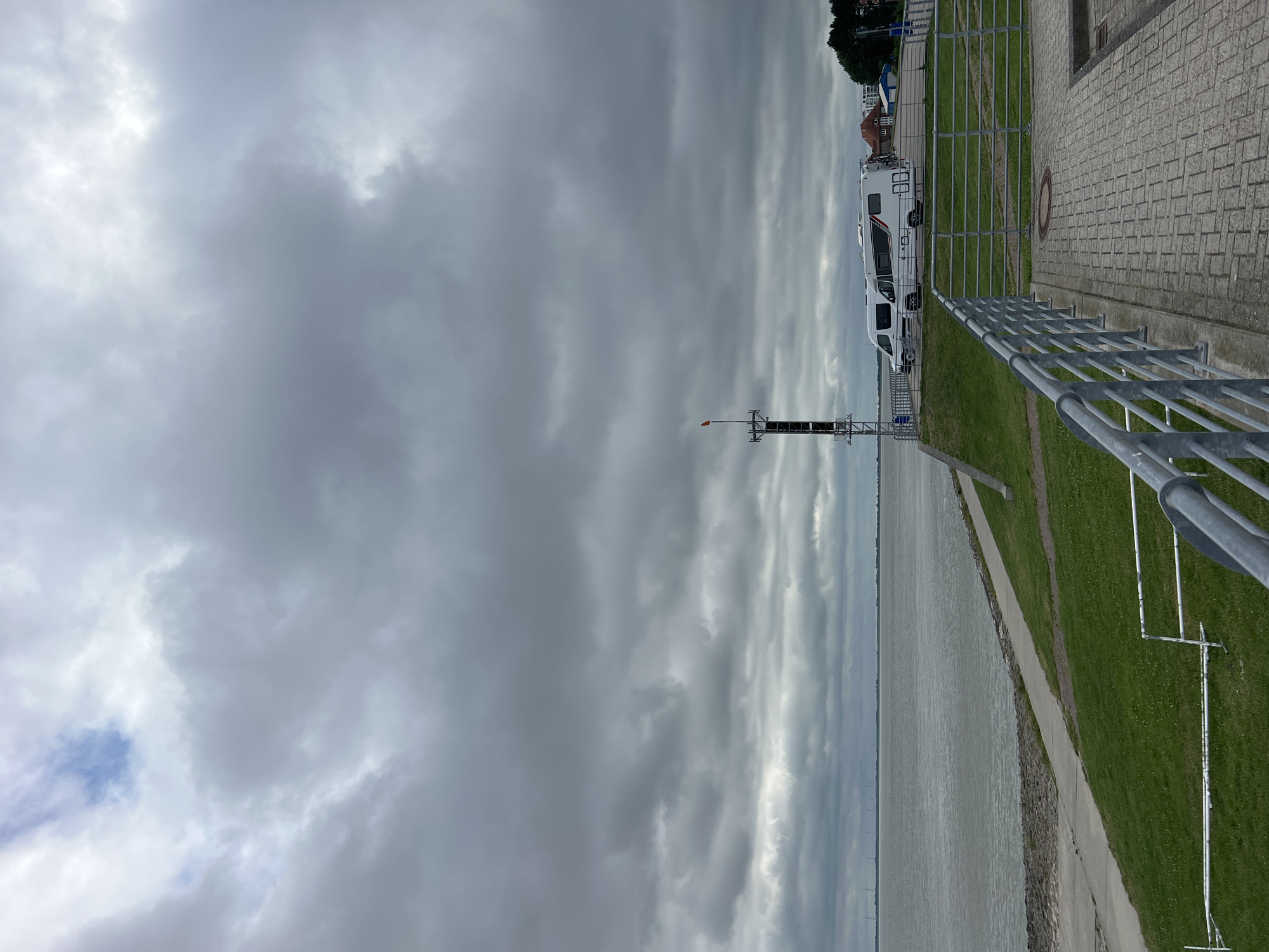 The image captures a cloudy day at the Jade Bay in Wilhelmshaven, Germany. In the foreground, a paved walkway with a metal railing runs diagonally across the frame. To the left, a grassy embankment slopes down towards the water. A concrete path is visible on the embankment. The water is calm and stretches to the horizon, where a line of distant wind turbines can be seen. On the raised area behind the embankment, two white camper vans are parked. A tall communication tower with a small orange flag at its top stands near the camper vans. In the background, buildings and trees are visible on the far shore.