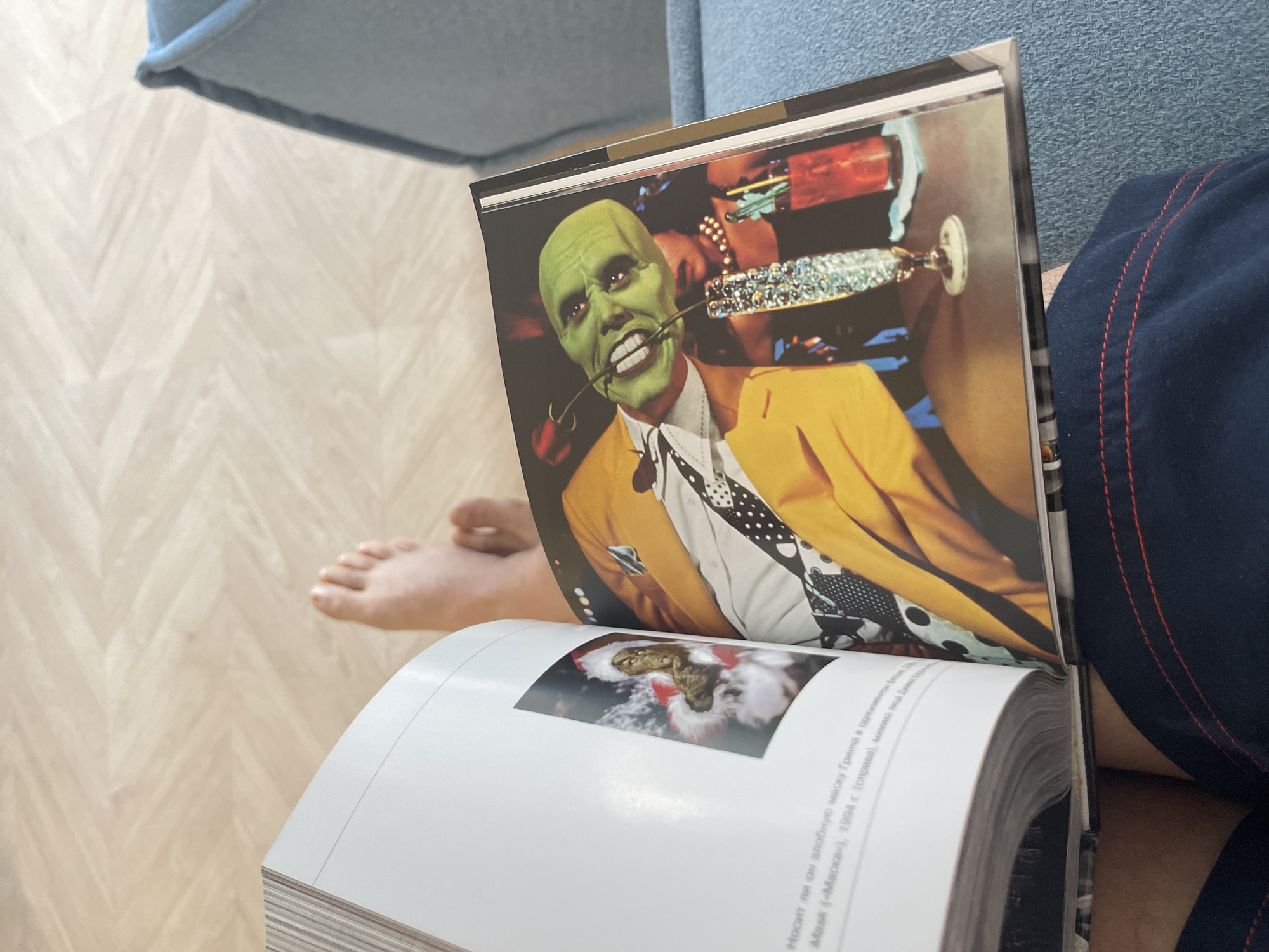 The scene depicts a book open, displaying an image of the character "The Mask" from the 1994 film, holding a rose in his teeth. A foot is visible in the periphery, and a person wearing dark blue shorts sits next to a blue couch. The location is Unknown, Unknown.