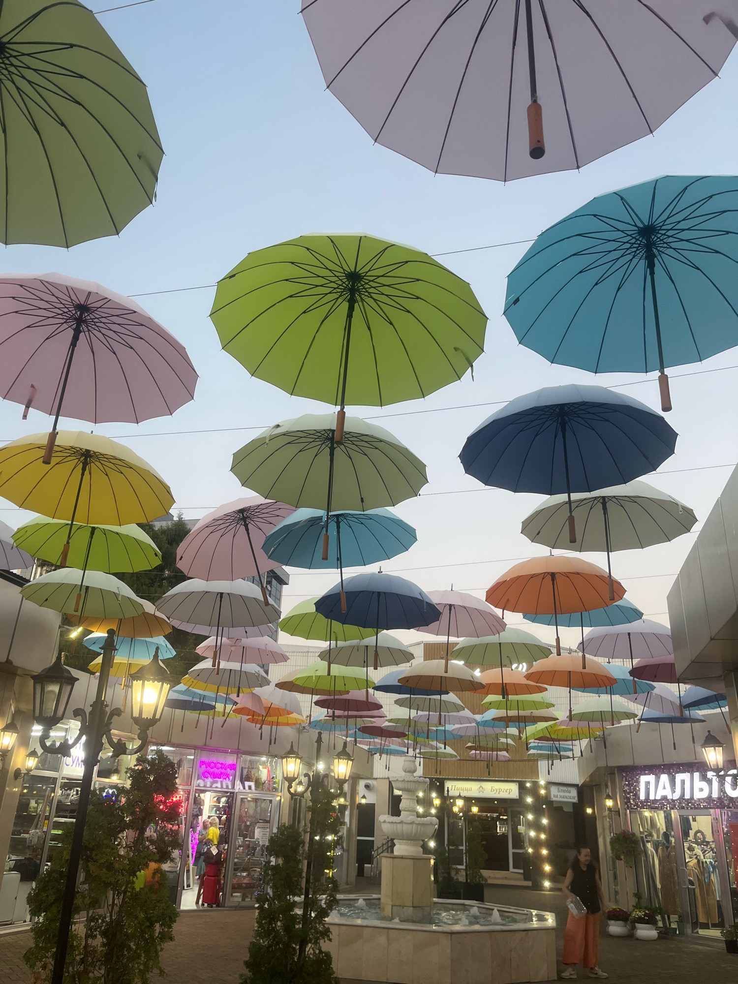 The image depicts an outdoor pedestrian walkway adorned with a canopy of colorful umbrellas suspended overhead. The umbrellas, in shades of pink, yellow, light green, light blue, and darker blue, are spread across the sky, creating a vibrant visual effect. Below the umbrellas, on either side of the walkway, are storefronts with illuminated signs. One sign reads "ПИЦЦА БУРГЕР", indicating a food establishment. Another, larger sign in purple lettering appears to say "ПАЛЬТО", suggesting a clothing store. Streetlights with glowing bulbs are positioned along the walkway, casting a warm light. In the foreground, a tiered fountain is visible. A person wearing an orange-pink outfit is walking in the middle ground, partially illuminated by the streetlights. The overall scene suggests a lively commercial or entertainment area. This image was taken in Sochi, Russia.