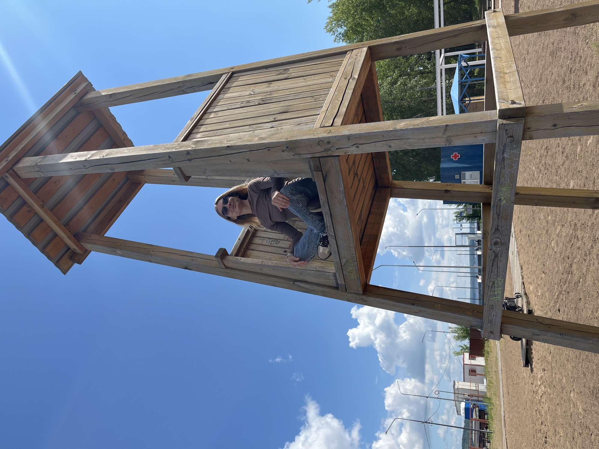 A young woman is captured sitting in a rustic, open-air wooden structure on a bright, sunny day in Chaikovsky, Russia. The structure, appearing to be an observation deck, lifeguard stand, or a large playhouse, is constructed from sturdy, weathered timber beams and planks. The woman, wearing a brown long-sleeved top, blue jeans, and light-colored sneakers, is looking directly at the camera from inside the structure, with dark sunglasses obscuring her eyes. Her hand, adorned with a ring, rests near her knee.
The sky overhead is a clear, vibrant blue with scattered white cumulus clouds. A strong sunbeam creates a lens flare in the upper left of the image. The ground around the structure is sandy or dirt, suggesting a beach or riverside setting, consistent with Chaikovsky's location near the Kama River/reservoir.
In the background, through the framework of the wooden structure, distant features are visible. A prominent blue building with a red cross symbol on its side stands out, possibly indicating a medical or first aid station. Beyond that, green trees line the horizon. Further in the distance, a body of water is discernible, dotted with light posts along its edge. Several street lights also extend into the mid-ground. A black, wheeled object, possibly a baby stroller, rests on the ground near the base of the wooden structure. Graffiti, reading "SKYL" or similar, is etched onto one of the wooden beams. The overall scene suggests a pleasant outdoor recreational area during daytime.
