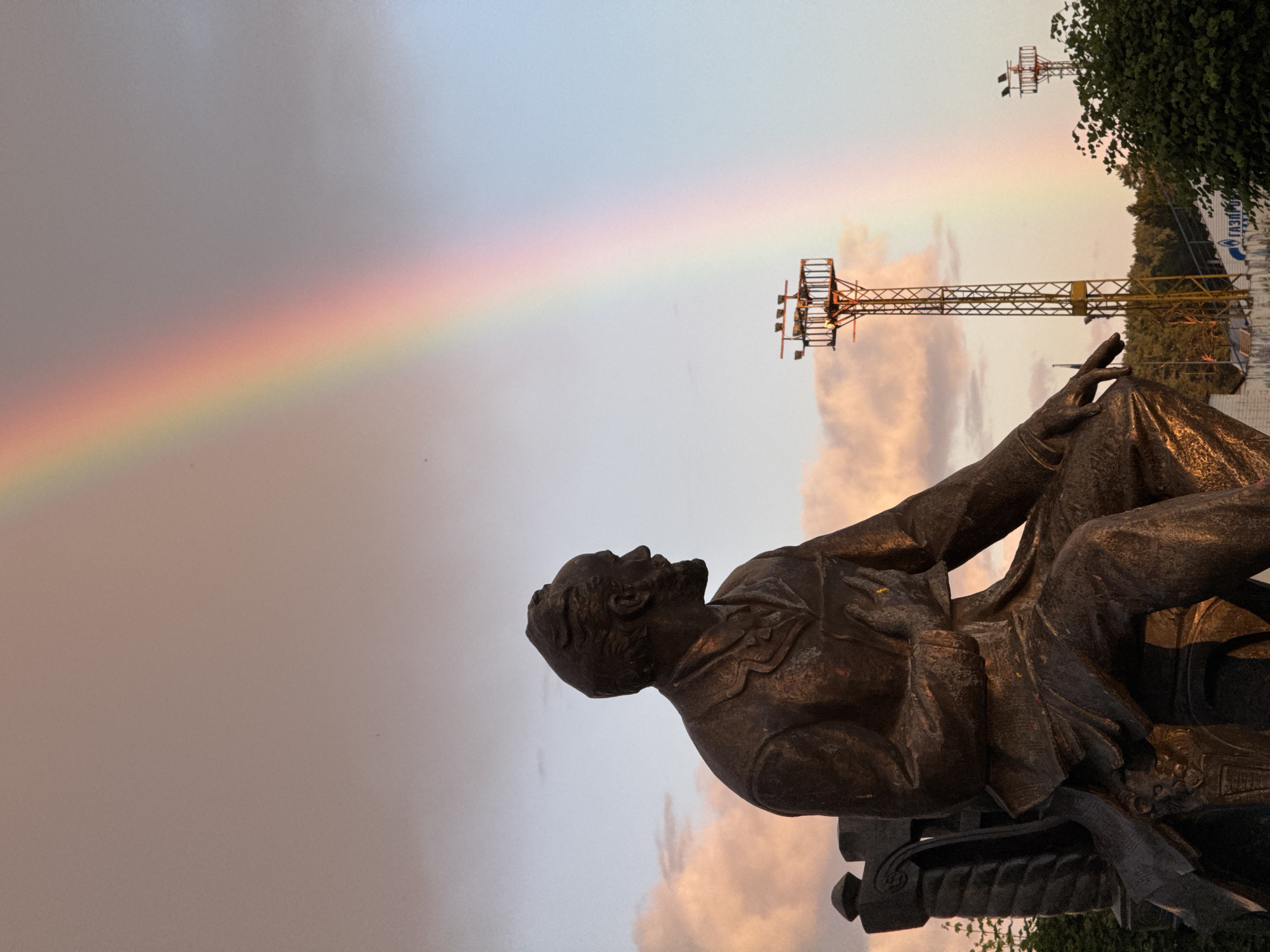 The image features a bronze statue of a seated man, possibly a historical figure, depicted in a contemplative pose. The statue is positioned in the foreground, with its metallic surface catching the light. In the background, a rainbow arcs across a sky that is transitioning from a hazy grey at the bottom to a lighter, almost pastel blue at the top. The sky is also dotted with soft, diffused clouds that are tinged with the warm colors of sunset or sunrise, suggesting it is either early morning or late evening.
Two industrial-looking towers with lighting fixtures are visible in the middle ground, one closer and taller than the other. Beyond these structures, a line of trees and foliage forms a backdrop, with some darker shadows suggesting the presence of other structures or a denser part of the landscape. On the right side of the frame, a portion of a white structure with blue lettering is visible, but the text is largely illegible due to distance and angle, although "ГАЗПРОМ" can be partially discerned.
The overall mood of the image is serene and slightly melancholic, with the dramatic natural phenomenon of the rainbow juxtaposed against the man-made elements of the statue and industrial towers. The soft lighting and atmospheric conditions contribute to a peaceful, yet perhaps poignant, atmosphere. The orientation of the photograph is vertical, with the statue appearing to be in the lower right quadrant when viewed in a typical landscape orientation.