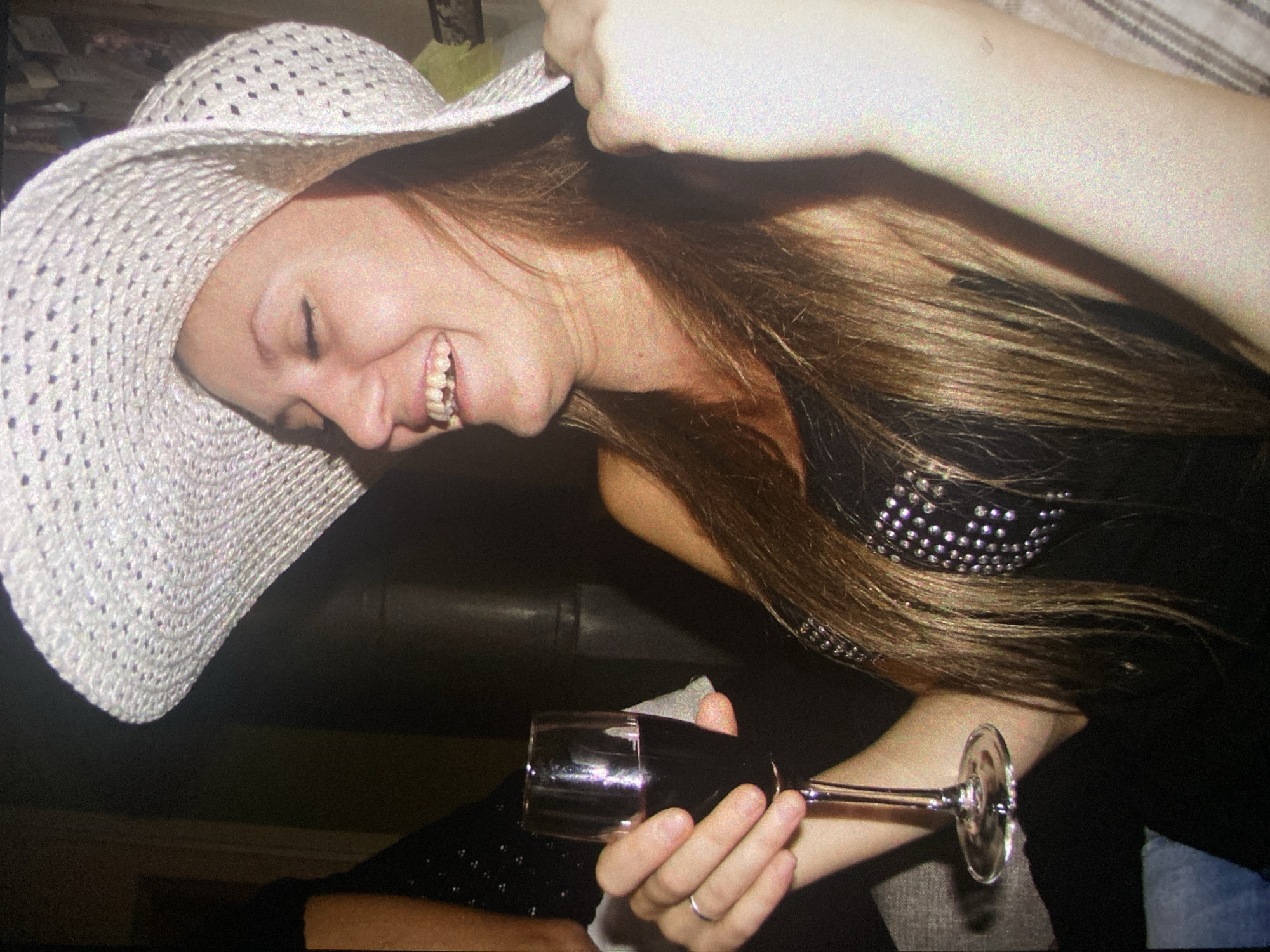 A woman with long brown hair, wearing a large white woven hat and a black top with silver embellishments, is smiling and holding a glass of red wine. She is also wearing a silver ring on her finger. The background is dark and partially obscured, but what can be seen appears to be a leather sofa. There is no indication of the location being Saint Petersburg, Russia.