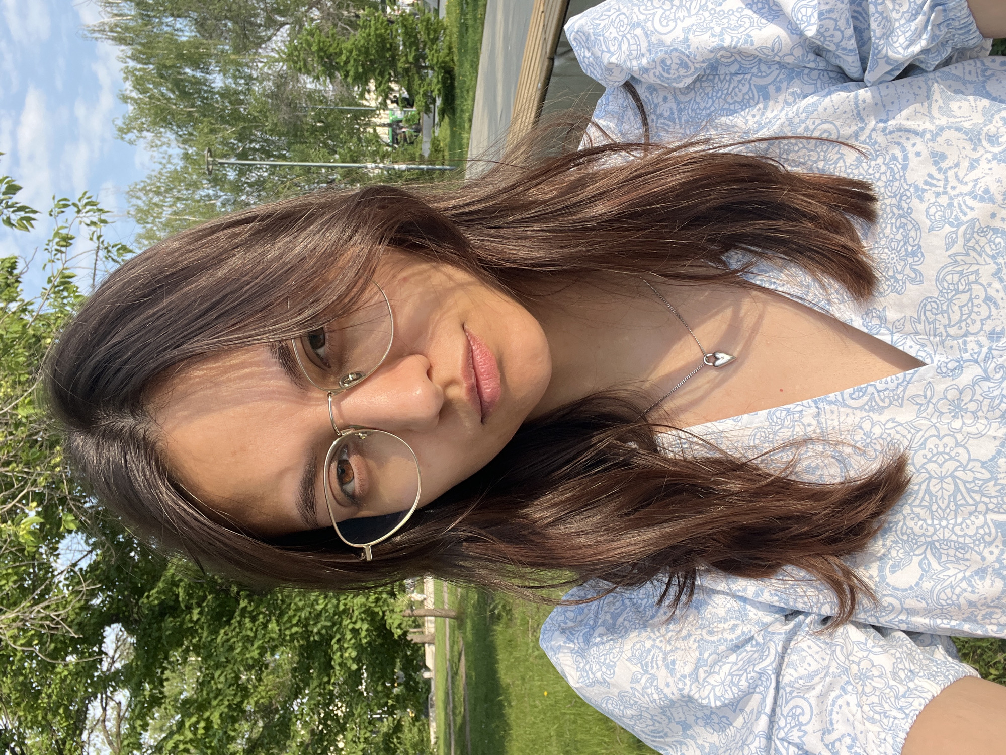A medium close-up shot of a woman wearing eyeglasses and a light blue printed top with a V-neckline. She also has a silver necklace with a pendant. The background shows a green park area with trees and grass, typical of an outdoor environment. This location has similarities to the city of Yekaterinburg, Russia.