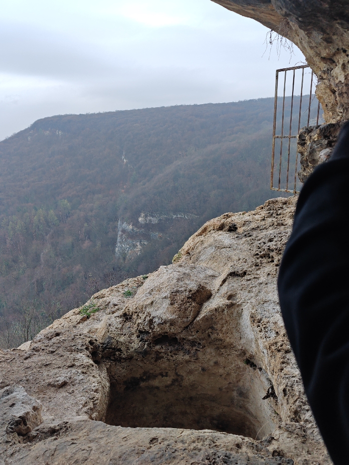 The scene depicts a rock-cut structure on the Shumen Plateau, Shumen, Bulgaria, under an overcast, grey sky. The background reveals a wide expanse of densely forested hills, exhibiting muted green and brown tones indicative of a non-foliage season, with sections of exposed rock cliffs. In the mid-ground, a rusty metal gate, composed of vertical and horizontal bars, is affixed to the rock face beneath an overhanging rock ceiling on the right side. The foreground features prominent, eroded light-colored rock formations. A distinct, rectangular, man-made cavity is carved into the rocky ground in the lower-center foreground. A partial view of a dark-clothed arm or shoulder is visible on the far right, suggesting an observer's perspective. Sparse dry vegetation hangs from the rock overhang, and small green plants adhere to the rocky surfaces.