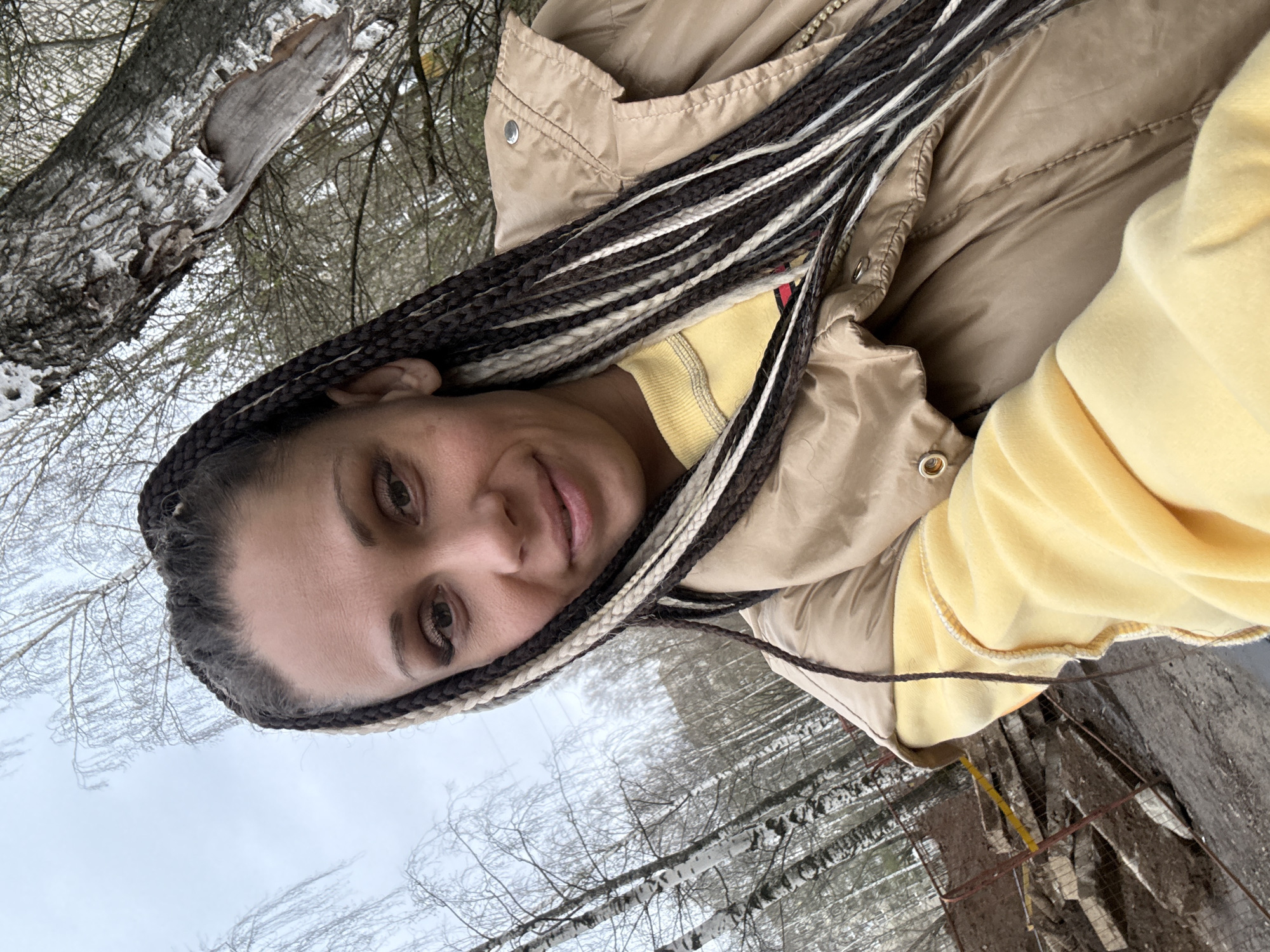 A selfie captures a woman outdoors in Rusanovo, Russia, amidst a wintry or early spring landscape. The woman, positioned prominently in the foreground and framed at an angle, looks directly at the camera with a soft, neutral expression. She has dark eyes and features, with long micro braids incorporating dark brown, light brown, and white strands. She is dressed warmly in a light tan or beige puffer vest, layered over a vibrant yellow long-sleeved top or hoodie, suggesting cold weather.
The background reveals a natural, wooded environment. Numerous bare trees with intricate branches dominate the scene, some exhibiting the distinctive white bark of birch trees. Patches of snow or frost are visible on the tree trunks and branches, and likely on the ground, indicating chilly conditions. The sky is overcast and bright, casting a soft, diffused light across the scene. The overall atmosphere suggests a serene, cold day in a natural setting. No text is visible in the image.