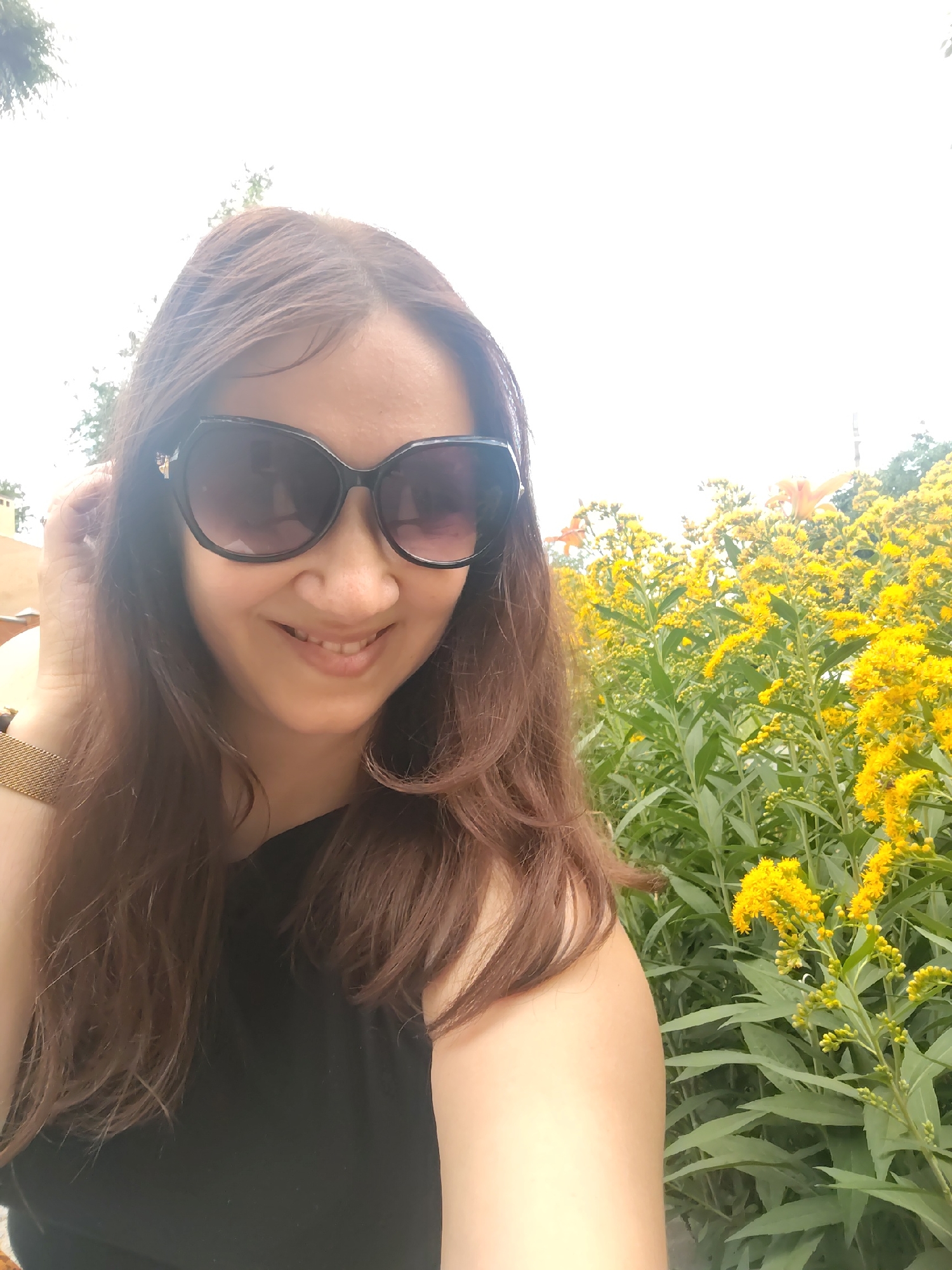 A woman is taking a selfie in front of a patch of tall, yellow wildflowers, possibly goldenrod. She has long, brown hair and is wearing large, dark sunglasses and a black top. A gold watch is visible on her left wrist. The background consists of lush green foliage and the bright sky. The location is identified as Rostov-on-Don, Russia.