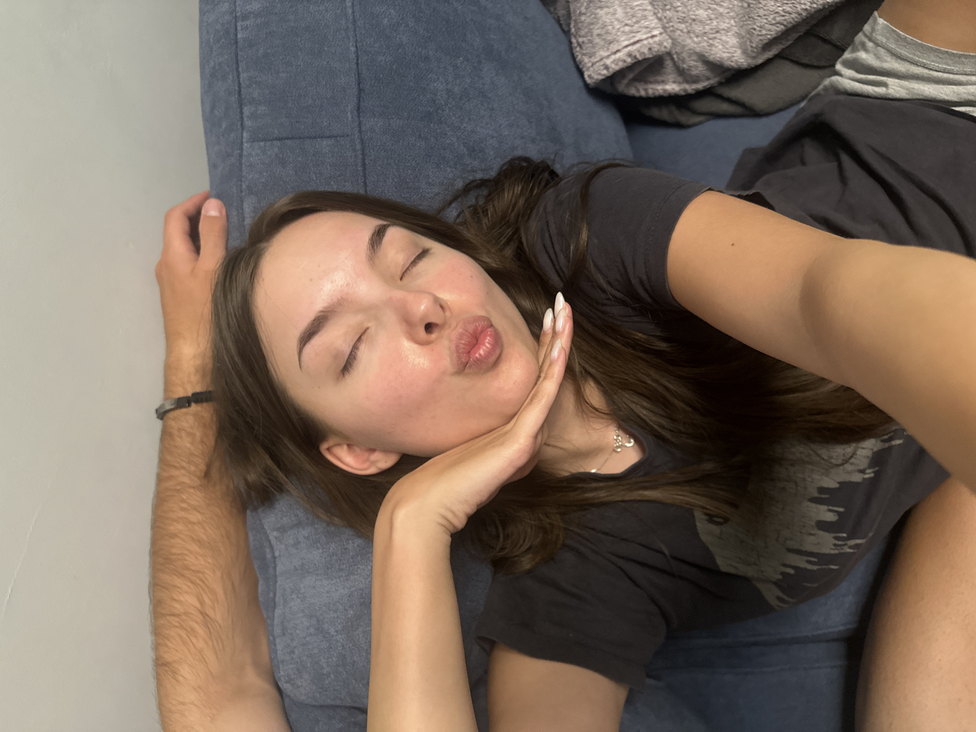 In this selfie, a young woman with long, dark hair and closed eyes is lying on a blue couch, puckering her lips as if to blow a kiss. Her right hand is raised, with her palm resting on her chin and her fingers gracefully curved. She is wearing a dark t-shirt with a graphic print. To the left of the frame, a man's arm, clad in a t-shirt and adorned with a bracelet, rests behind the woman's head. The background consists of a plain wall and a folded gray blanket on the couch. The scene suggests an intimate and relaxed setting, potentially a living room in a home.