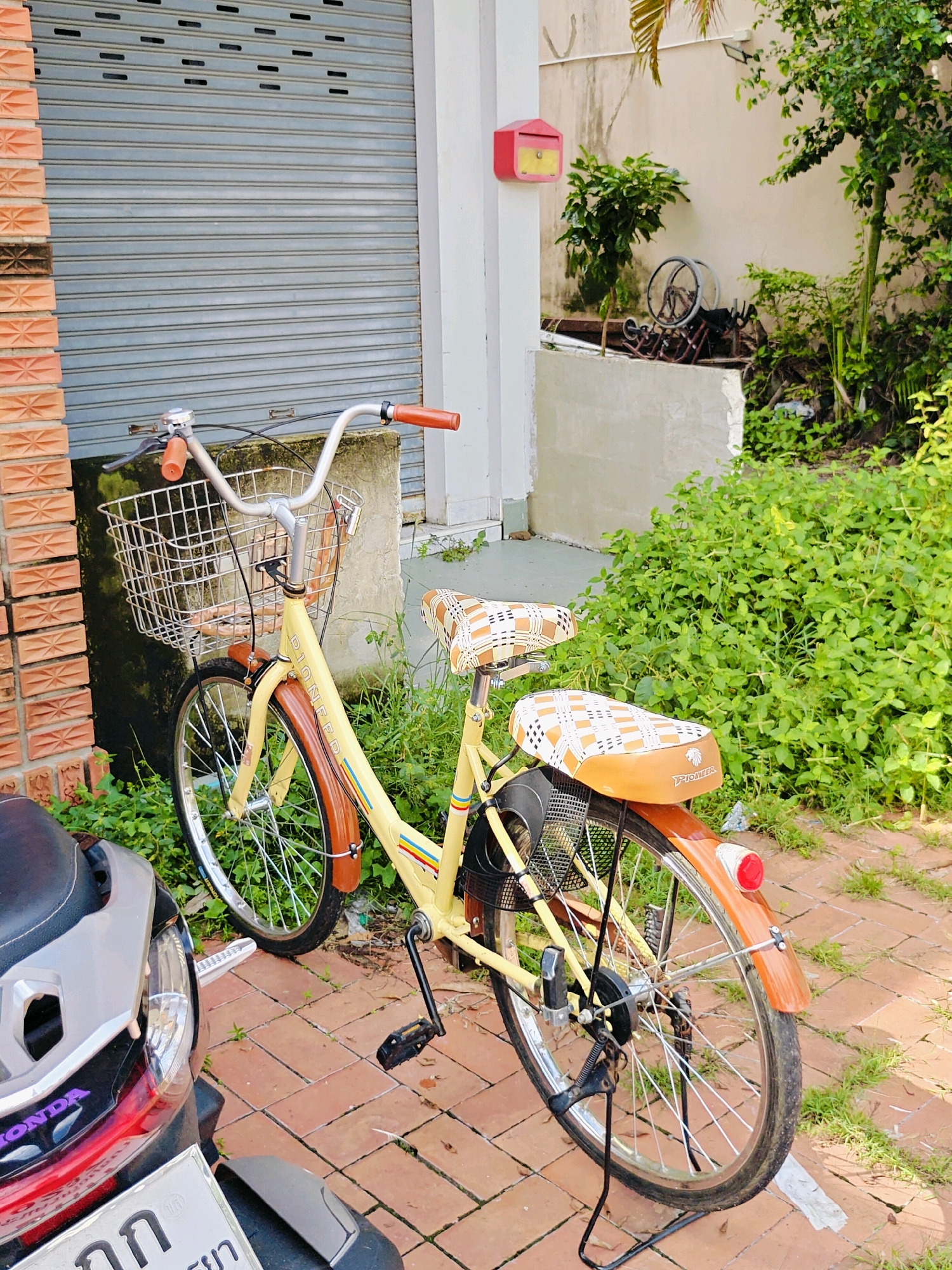 The image depicts a yellow and brown vintage bicycle parked on a brick pathway in front of a grey roller shutter door and a building. The bicycle has a silver wire basket on the handlebars, brown grips, and a brown and orange checkered seat. A dark-colored motorcycle is partially visible in the lower left corner. To the right of the bicycle is a dense patch of green vegetation. In the background, a red mailbox is mounted on the wall of the building. The overall scene suggests an outdoor setting within a residential or commercial area in Phra Nakhon Si Ayutthaya City Municipality, Thailand.