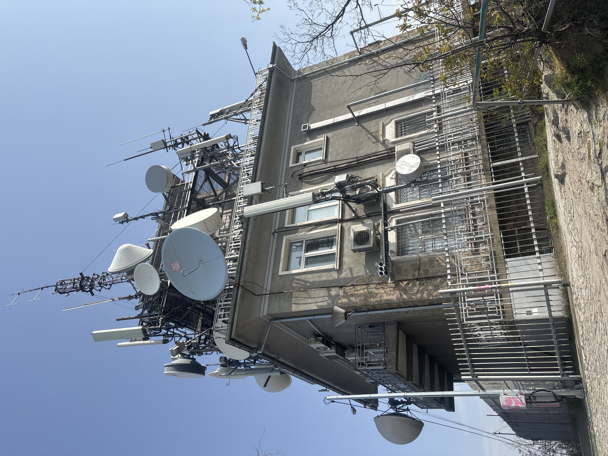 The image shows a building in Plovdiv, Bulgaria, heavily outfitted with telecommunications equipment. A large array of satellite dishes and antennas is mounted on a metal structure attached to the building's side. The building itself appears to be a multi-story residential structure with a gray facade and windows. Air conditioning units are visible on the building's exterior. The sky is clear and blue, suggesting a sunny day. A concrete structure, possibly a retaining wall or a neighboring building, is visible to the right, partially obscured by foliage.