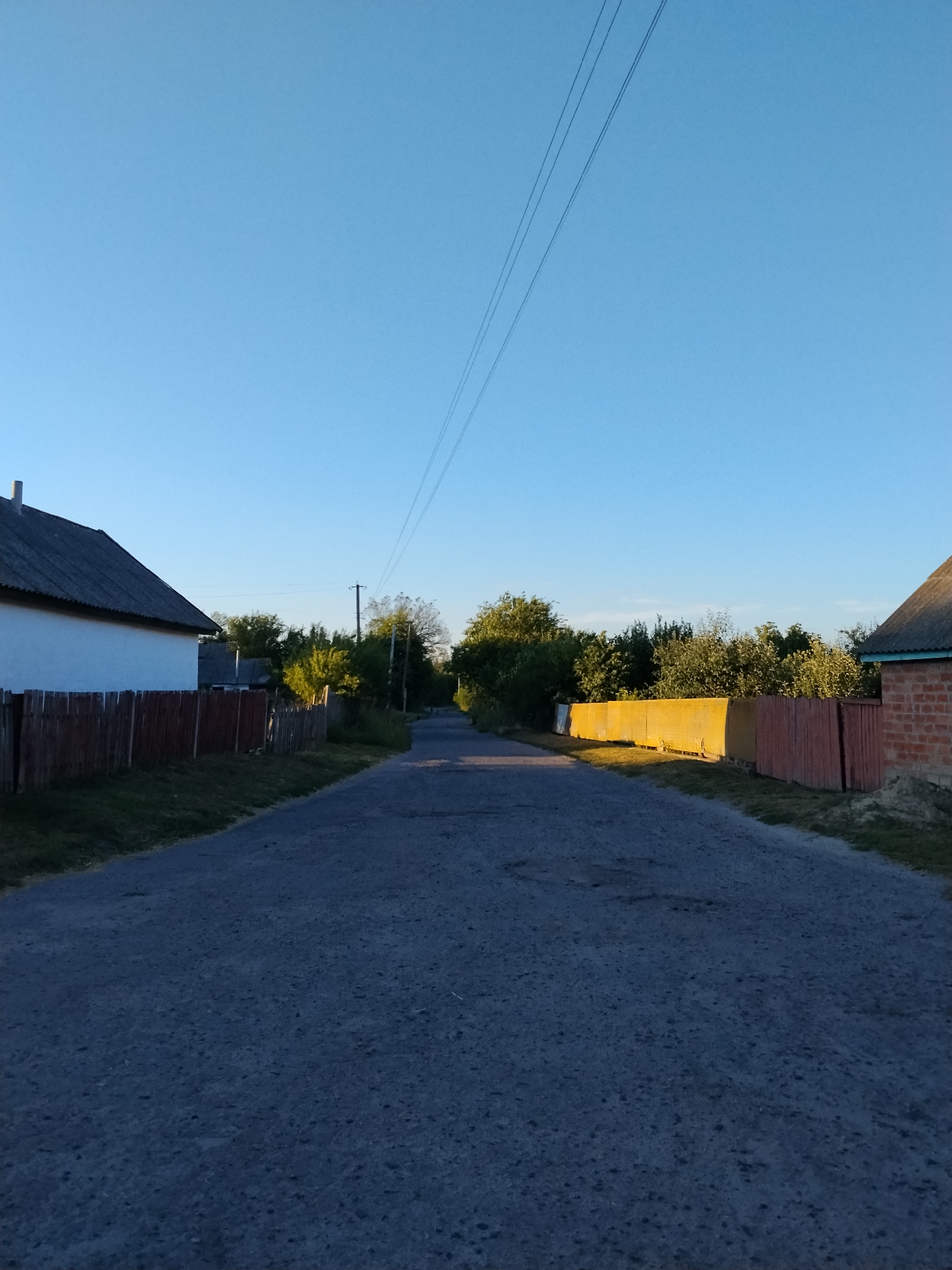 The image depicts a rural road in Ustyvytsia, Ukraine, under a clear blue sky. The road appears to be paved with asphalt and shows some signs of wear and tear. On the left side of the road, a white building with a dark roof and a wooden fence in front of it is visible. Further down the road on the left, more trees and buildings can be seen. On the right side of the road, there is a long, bright yellow fence followed by a red wooden fence and a brick building with a dark roof. Trees and foliage line both sides of the road, creating a sense of a village or countryside setting. Overhead, power lines stretch across the sky. The lighting suggests it is either early morning or late afternoon, with the sun casting long shadows. There are no people or animals visible in the scene.