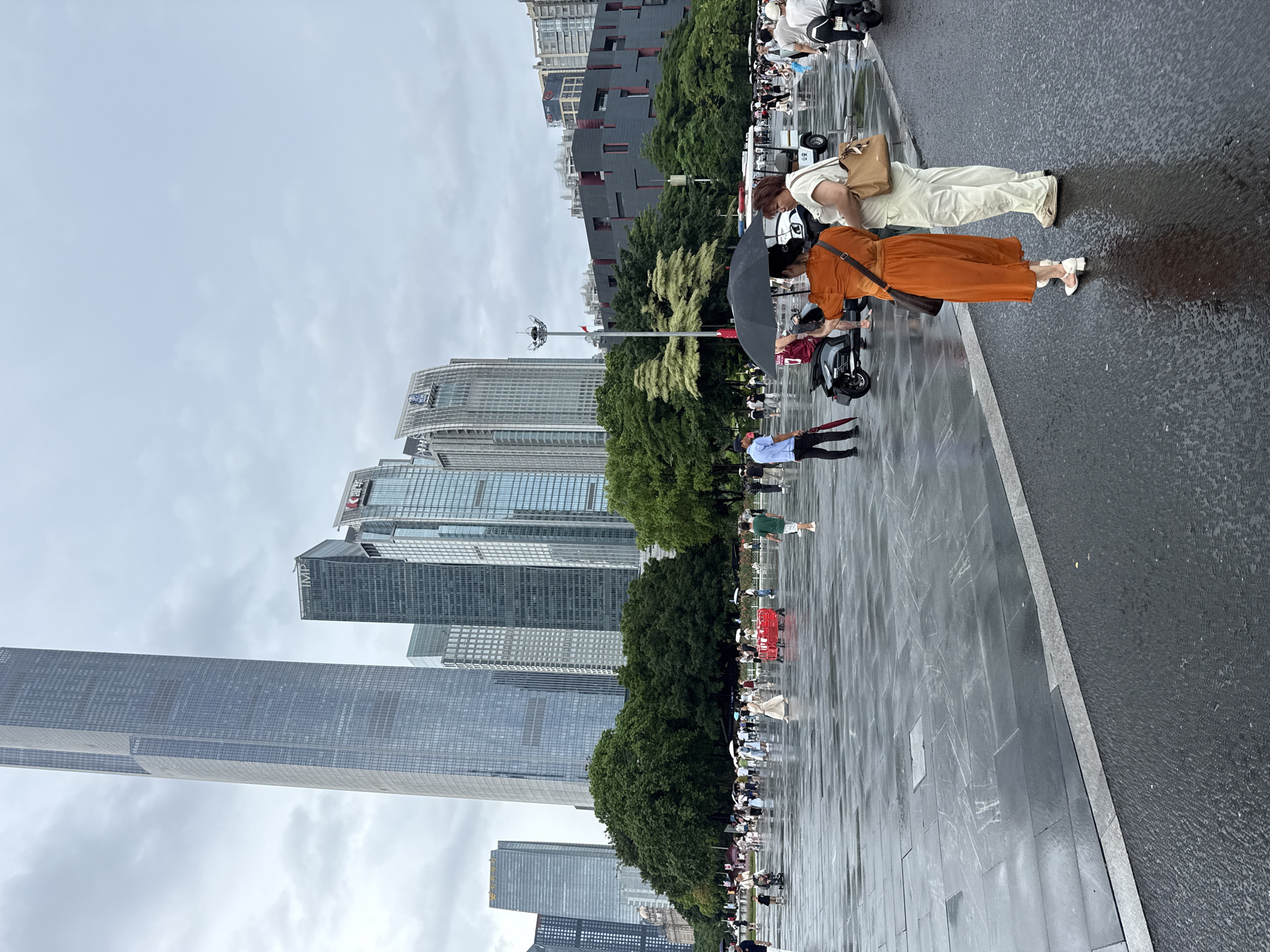 The image captures a wide-angle view of a wet, paved plaza in Tianhe District, China, under an overcast sky. Several modern skyscrapers dominate the background, suggesting an urban setting. In the foreground, two women are standing, facing away from the camera, with one holding a black umbrella over her head. The woman on the left wears an orange dress and the woman on the right wears a cream-colored outfit. They appear to be conversing or observing something together.
The plaza is populated with numerous other people walking, some with umbrellas, indicating it may be raining or has recently rained. A red cart is visible in the mid-ground, along with a few electric scooters or golf carts. Lush green trees line the perimeter of the plaza behind the crowd. The wet pavement reflects the light and the figures, contributing to the moody atmosphere. The overall scene suggests a public space in a bustling city, likely a popular gathering spot.