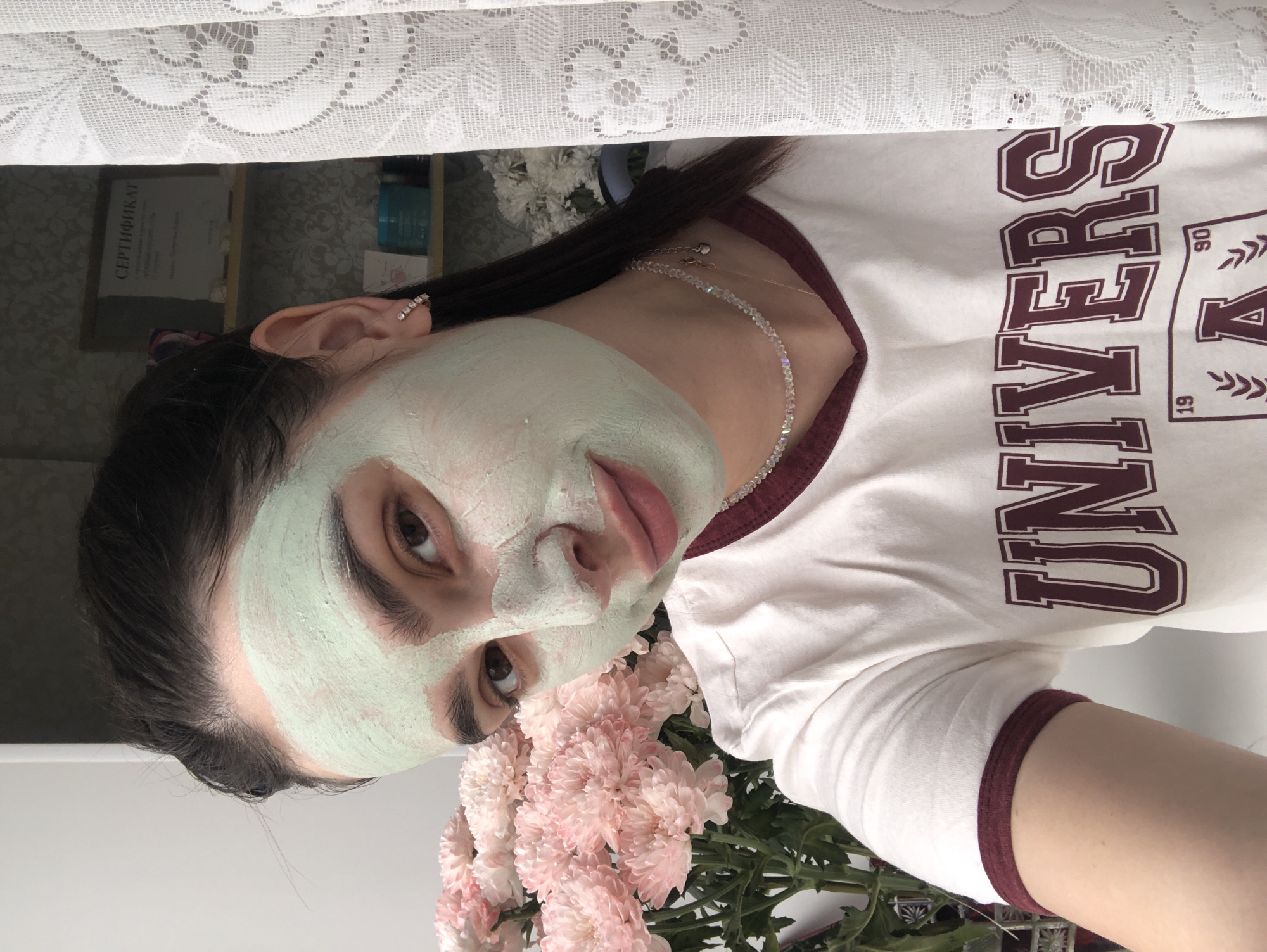 A young woman with dark hair is shown in this media file, applying a light green face mask. She is wearing a white t-shirt with maroon trim and text that reads "UNIVERSITY". She has on a delicate necklace and a ring on her finger. In the foreground, a bouquet of light pink chrysanthemums is visible. The background appears to be an indoor setting, with a patterned wallpaper and a white lace curtain. There is also a certificate or document visible in the background. The lighting suggests it could be daytime. The overall mood is relaxed and focused on self-care.
