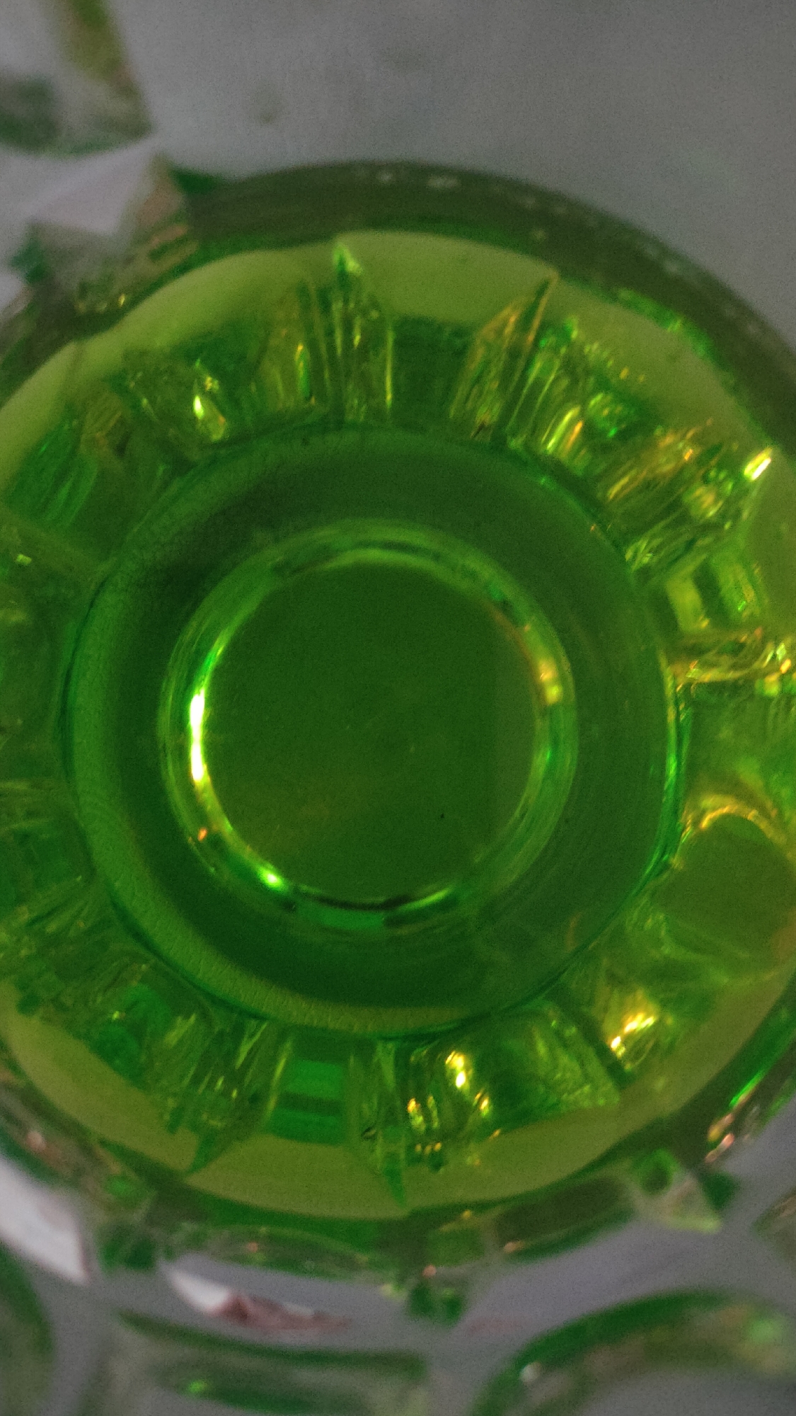 This is a close-up, top-down view of a green glass. The glass appears to be transparent with a textured rim. The lighting creates reflections and highlights on the surface of the glass, giving it a glossy appearance. There is no visible indication of location within the city of Bolotnoye, Russia. The image solely focuses on the glass object.