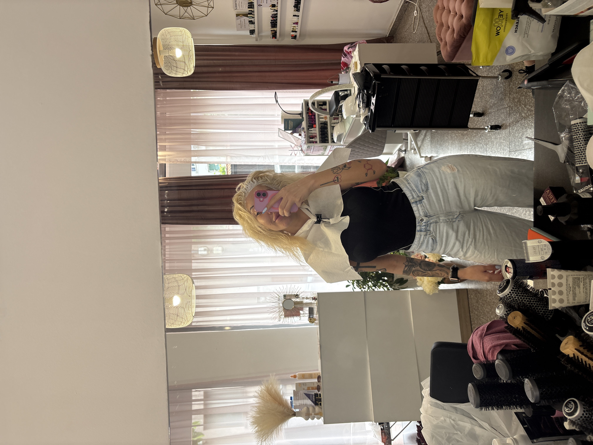 The primary image depicts a blonde woman taking a mirror selfie in what appears to be a modern hair or beauty salon, located in Plovdiv, Bulgaria. The woman is wearing a black top, light blue ripped jeans, and a white disposable cape or collar, suggesting she is mid-treatment or has recently finished. She holds a pink smartphone to capture her reflection, partially obscuring her face. Her arms display several tattoos, including a chemical structure and a heart containing the number '1'.
The salon is well-lit, with natural light entering through large windows covered by sheer and darker brown curtains. The decor includes light-colored walls, a ceiling-mounted geometric light fixture, and wall lamps with woven shades. Visible furniture includes a white cabinet adorned with a sunburst mirror and a vase of pampas grass, along with a plush pink chair and a black salon chair.
To the right, a black salon trolley is positioned near various hair care tools such as brushes and rollers. Shelves on a far wall display numerous bottles, likely nail polishes, interspersed with framed certificates, one of which clearly reads "ДИПЛОМА" (Diploma). Other visible objects include a yellow bag with "WOOLRY" and "AEON" printed on it, a can labeled "CELANO," and product bottles partially identifiable as "OLAPLEX" and "AZURE." The overall atmosphere suggests an active and functional professional salon environment.