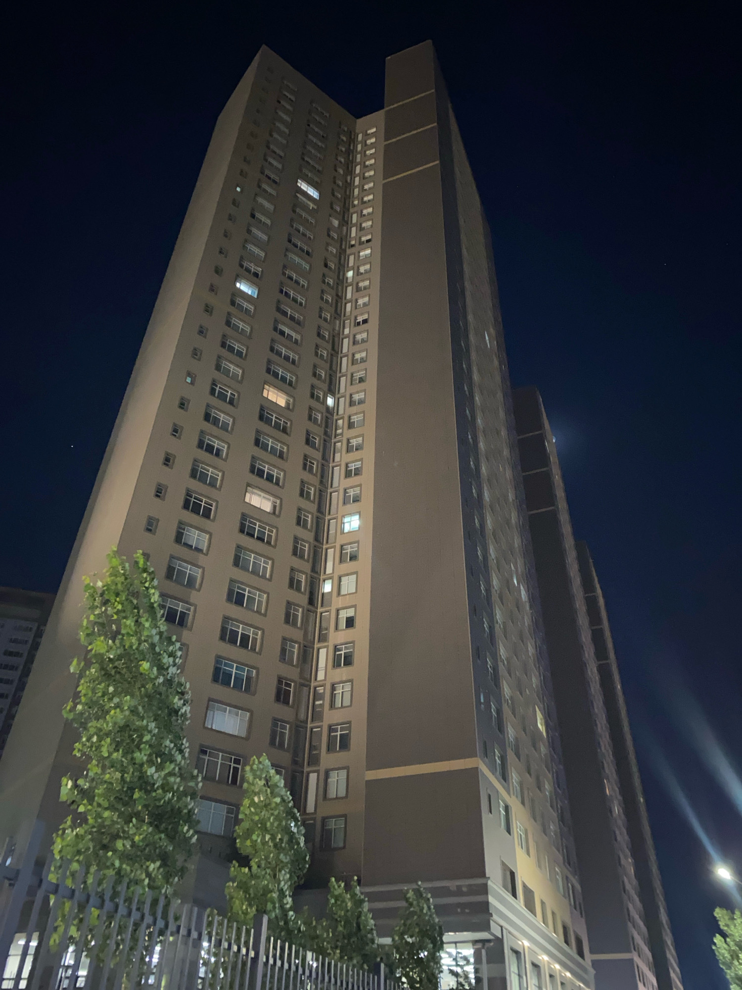 The image shows a view from below of two very tall, modern apartment buildings at night. The buildings are dark gray with numerous windows, some of which are lit from within. The sky above is a deep blue-black, with a few faint stars visible. In the foreground, there are several trees with green foliage, illuminated by ambient light. A metal fence runs along the bottom of the image, separating the viewer from the trees and buildings. The lighting suggests the presence of streetlights or lighting from within the buildings. The architectural style of the buildings, their scale, and the overall ambiance are consistent with a contemporary urban setting in Astana, Kazakhstan. There are no people visible in the image.