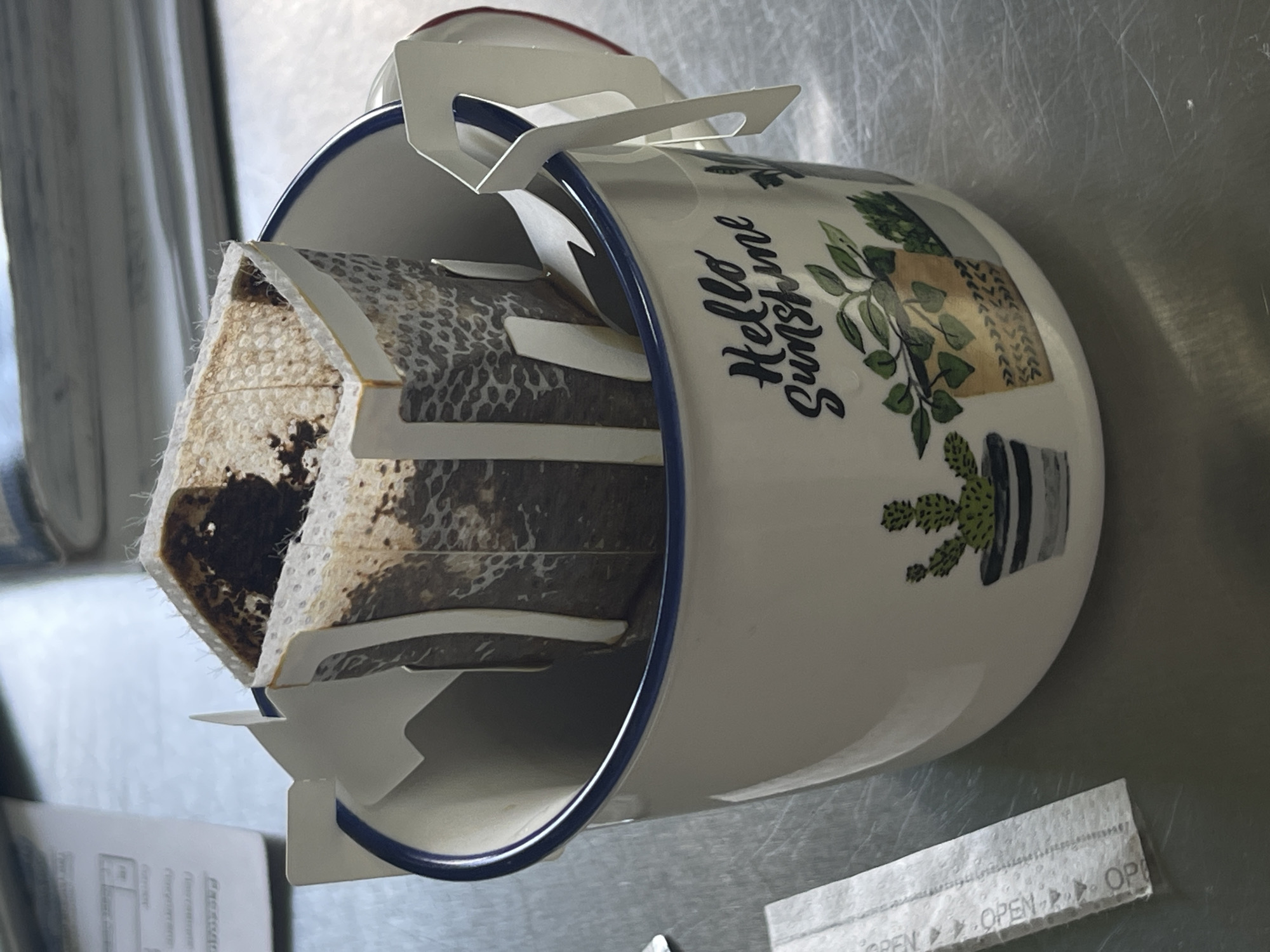 The image shows a white ceramic mug with a blue rim, decorated with cartoon-style potted plants and the words "Hello Sunshine" in a whimsical font. Inside the mug, a paper coffee filter bag filled with coffee grounds is suspended by white cardboard hangers that hook over the rim of the mug. The coffee filter appears to be used and slightly damp, suggesting that coffee has recently been brewed or is in the process of brewing. The mug rests on a metallic, possibly stainless steel, surface, which appears to be a countertop or table. In the foreground, to the right of the mug, a small white paper strip with the word "OPEN" repeated is visible. The lighting suggests an indoor setting, with some ambient light sources reflecting off the metallic surface. The overall scene evokes a sense of a morning coffee ritual or a moment of relaxation. There are no people visible in the image.