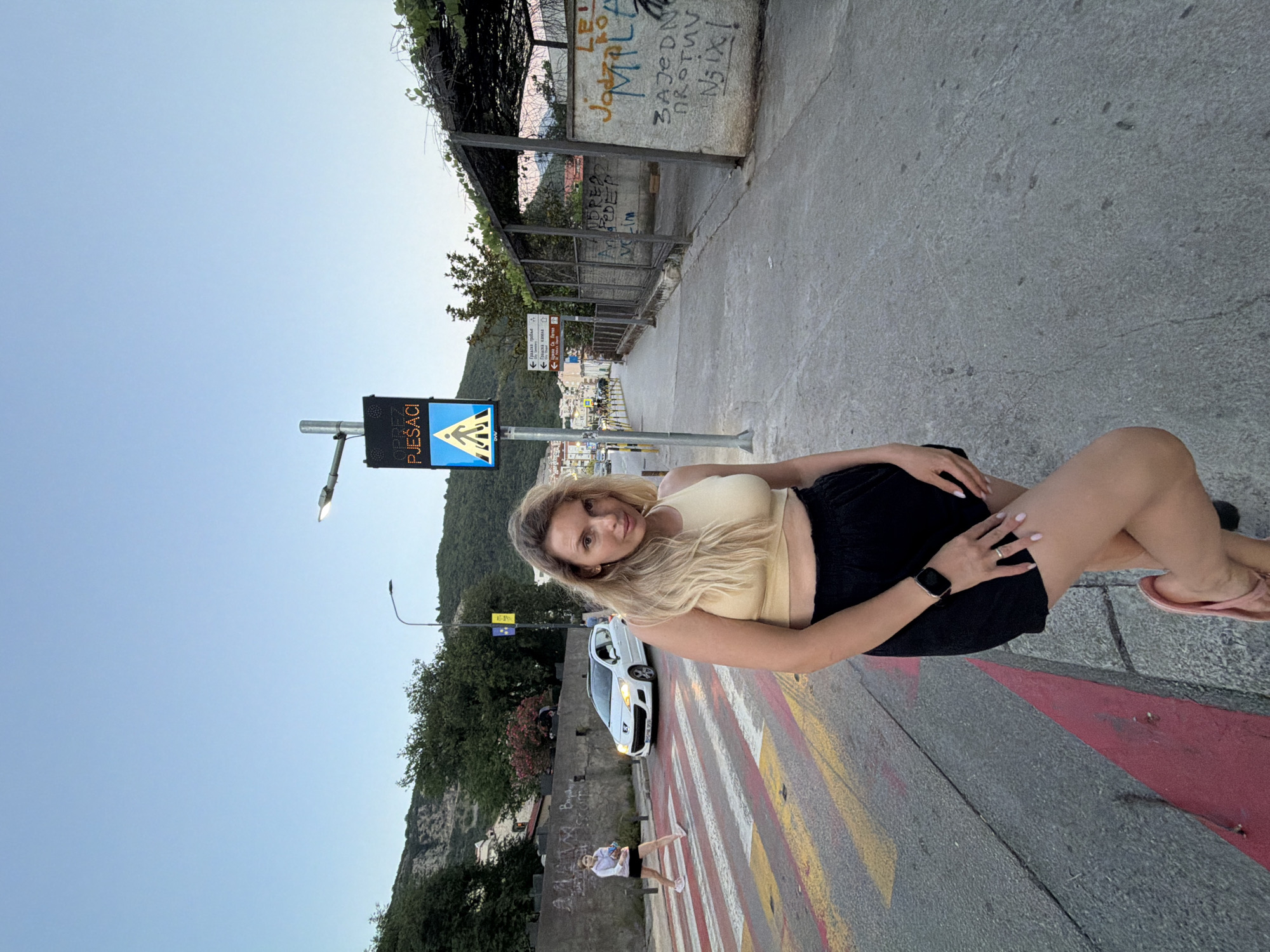In Budva, Montenegro, on a clear day with a soft twilight sky, a woman with blonde hair stands on a pedestrian crossing. She is wearing a yellow crop top and black shorts, with her left leg bent. The pedestrian crossing has red and yellow stripes. Behind her, a white car is driving on the road, and another person is walking on the sidewalk. A pedestrian crossing sign with a walking person icon and text is visible. The environment includes trees, a concrete wall with graffiti, and a wire fence.