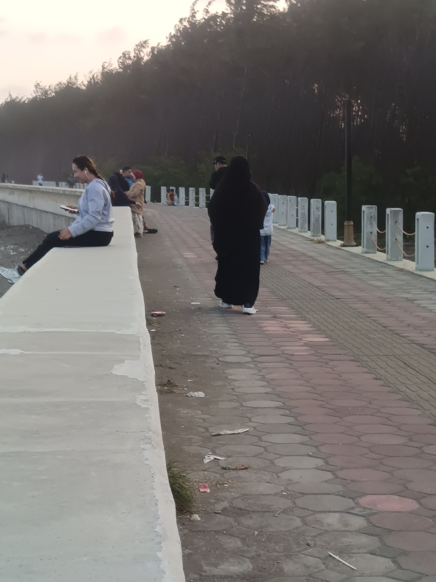 The image depicts a walkway or promenade in Kelurahan Tegalkamulyan, Cilacap, Indonesia, likely near the coast due to the presence of trees and a sandy/rocky area. The walkway is paved with hexagonal bricks in shades of red and gray. A low, concrete barrier runs along the left side of the walkway, with a woman in a grey hooded sweatshirt and black pants sitting on it, looking at her phone and wearing earbuds. Further down the walkway, a person in a black abaya and white shoes is walking away from the camera. Other individuals are visible in the background, including a person in a tan jacket and red headscarf, and a child. A line of decorative posts with ropes runs along the right side of the walkway. The environment appears to be outdoors, with a tree line visible in the distance under a hazy sky, suggesting either early morning or late afternoon light. Some debris, including paper and plastic, is scattered on the ground near the walkway.