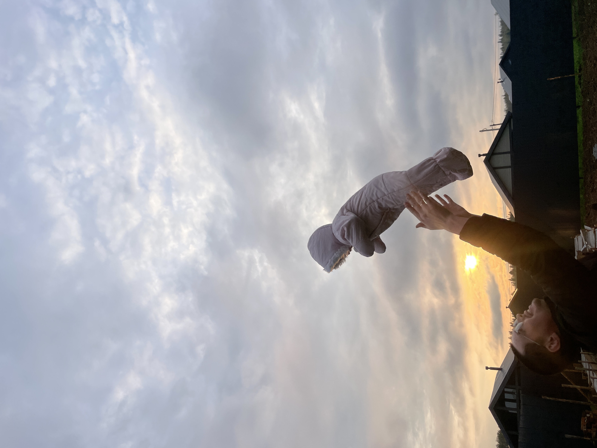 A person is throwing a grey baby snowsuit into the air against a cloudy sky. The setting appears to be outdoors with houses and trees in the background. The sun is visible through the clouds. The location is Подшивалово, Russia.
