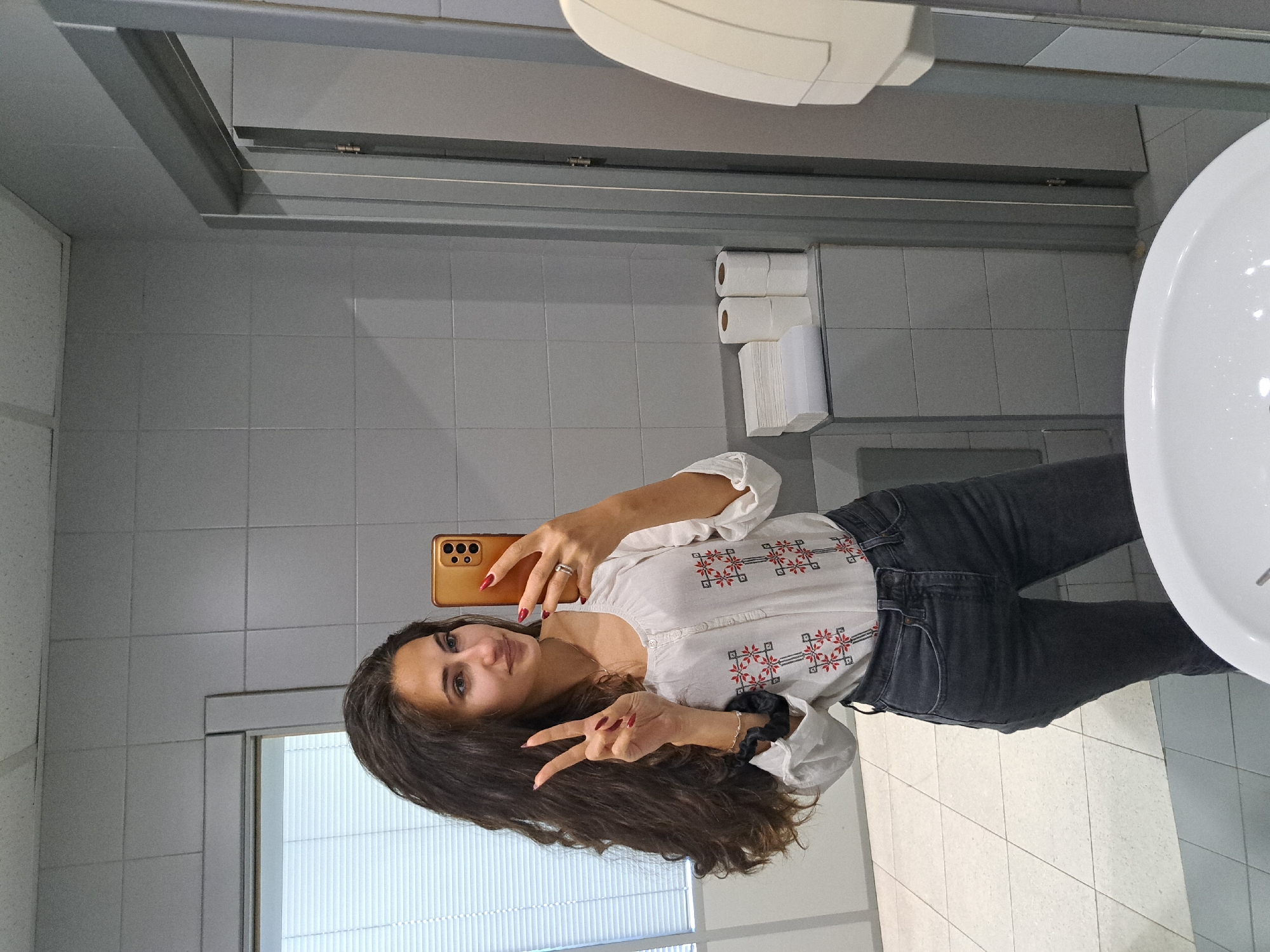 The image shows a young woman with long, dark hair taking a selfie in a public bathroom. She is wearing a white embroidered blouse and dark jeans, and making a peace sign with her fingers. She is holding a gold-colored smartphone.
The bathroom has grey tiled walls and floor. A white sink and hand dryer are visible. There is also a toilet paper dispenser. The background shows what appears to be a window with blinds.
The image does not provide information about the location of the bathroom in Sofia, Bulgaria.