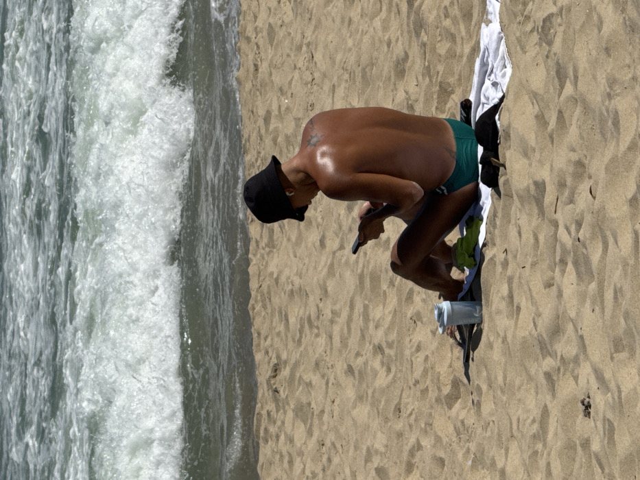A shirtless person wearing a black bucket hat and green swim trunks stands on a sandy beach near breaking waves. The individual bends over, looking at a cell phone in their hand. A white and green towel and a blue water bottle sit nearby on the sand. The scene suggests a casual moment on the beach, perhaps checking messages or taking photos.