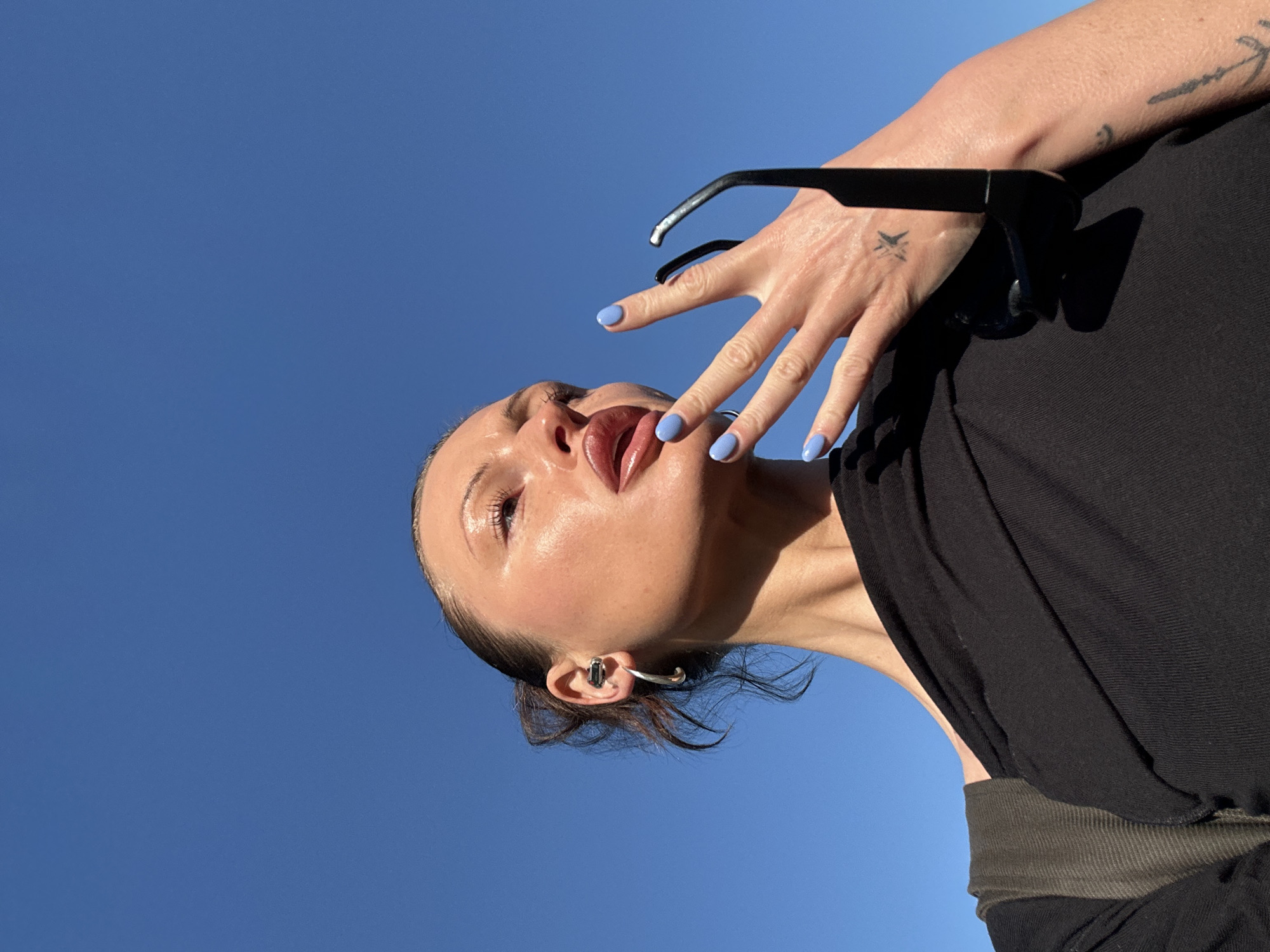 A low-angle portrait captures a person's upper body against a clear, bright blue sky. The subject's head is tilted significantly to the left, looking upwards with slightly parted lips. Their skin has a luminous, dewy appearance, reflecting the strong sunlight. A hand, adorned with light blue nail polish, is raised near the face, holding a pair of black-framed sunglasses. A small, stylized tattoo resembling crossed lines or an 'X' is visible on the back of this hand, with another indistinct tattoo further up the forearm. The person is wearing a black garment, and a distinct silver-toned ear cuff or earring is visible on their right ear. The brilliant sunshine creates crisp shadows and highlights, emphasizing the contours of the face and hand. The background is an expansive, cloudless blue sky, suggesting a sunny daytime scene. The activity appears to be posing for a photograph. The setting is Tunbridge Wells, United Kingdom.