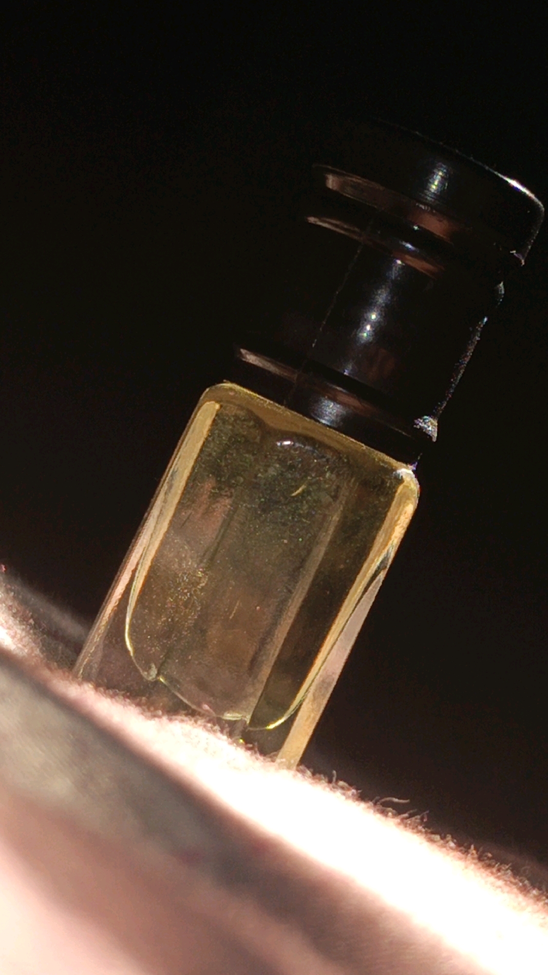 A macro photograph captures a small, rectangular glass vial filled with a yellowish liquid, possibly an oil or perfume. The vial has a dark brown, ridged cap. The vial is positioned at an angle, resting on a textured, fibrous surface that appears to be a light-colored fabric. The background is dark and out of focus. The lighting creates highlights on the glass and cap, with a bright light source illuminating the fabric from the lower left. There are no people visible, and the scene is static. The image is a close-up, offering no contextual information about its location, including the city of Nabinagar, Bangladesh.