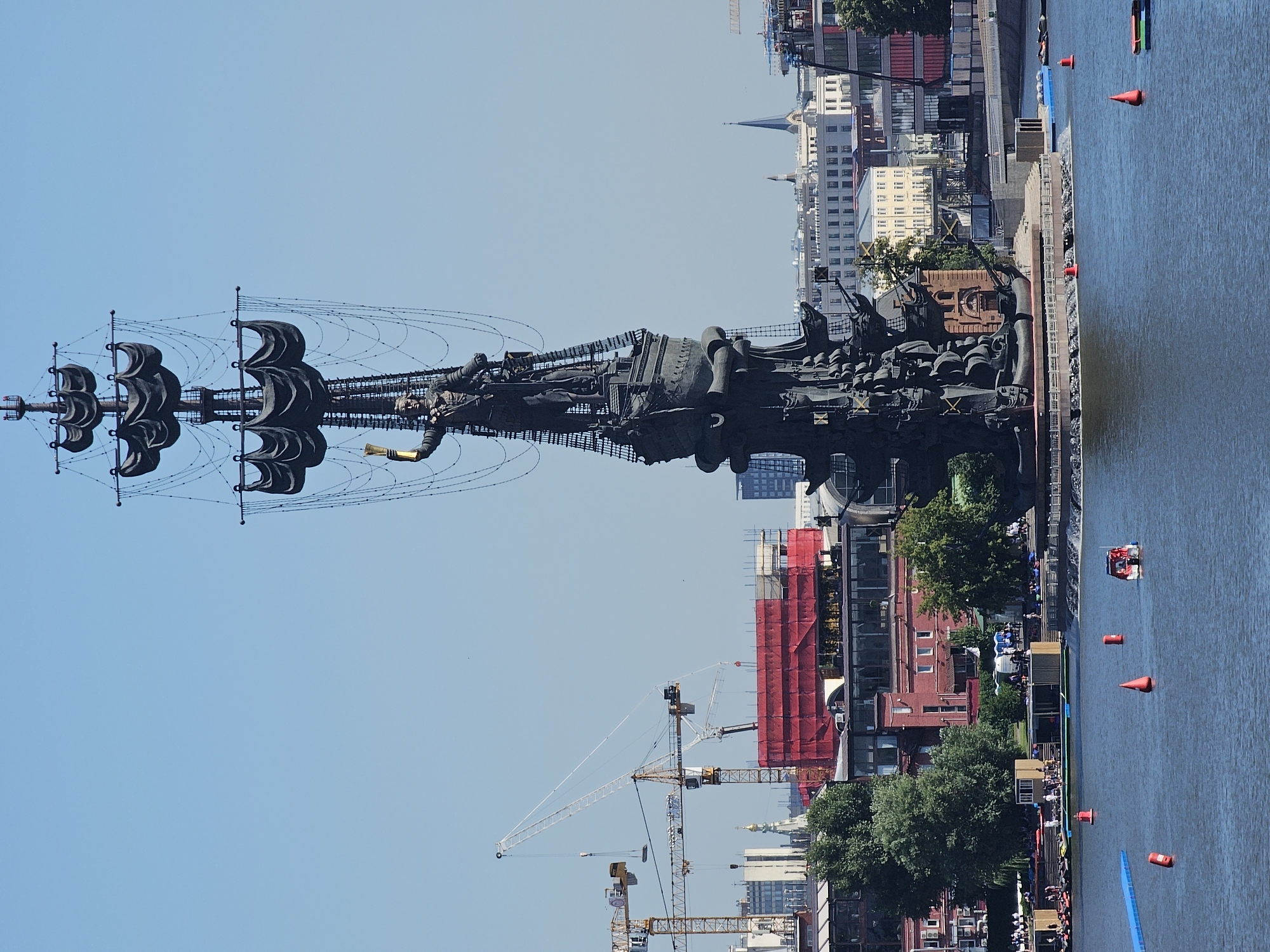 A monumental statue of Peter the Great, depicted as a captain on a ship's mast holding a golden staff, dominates the center of the frame. The statue is dark bronze and features intricate detailing on the ship's hull and sails, which are also dark. The statue stands on a circular stone base in the foreground, surrounded by water. In the background, a city skyline is visible, with various buildings, some with spires. Construction cranes are also present, suggesting ongoing development. The sky is clear and blue. In the water, several red buoys are scattered, and a small red and white boat with people is visible. On the shore to the left, there are trees, buildings with red brick facades, and people gathered. The overall scene depicts a large, imposing historical monument in an urban waterfront setting on a clear, sunny day.