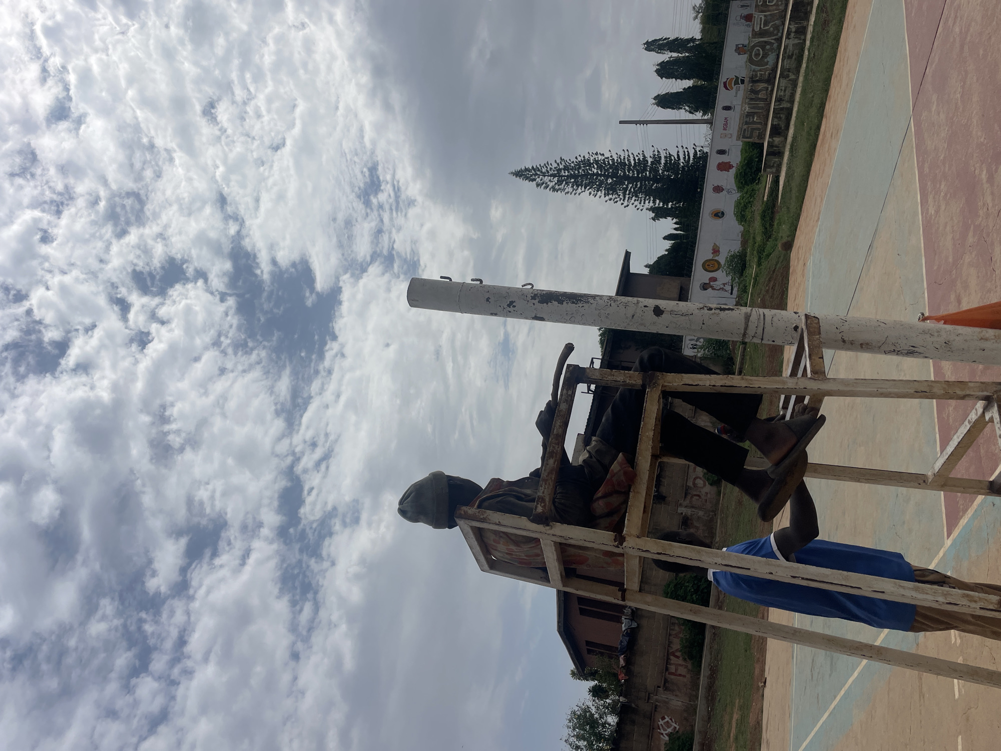 A street scene, likely in Gura Top, Nigeria, shows two individuals on a white metal structure, possibly a lifeguard stand or elevated platform. One person sits atop the structure, wearing dark clothing and a hat. Another person in a blue shirt stands partially obscured at the base of the structure. The background includes a painted wall with cartoonish figures and words, possibly related to sports. A pine tree and power lines are also visible. The sky is overcast with broken clouds. The ground appears to be a faded sports court with painted lines. A building is visible in the background behind the painted wall.