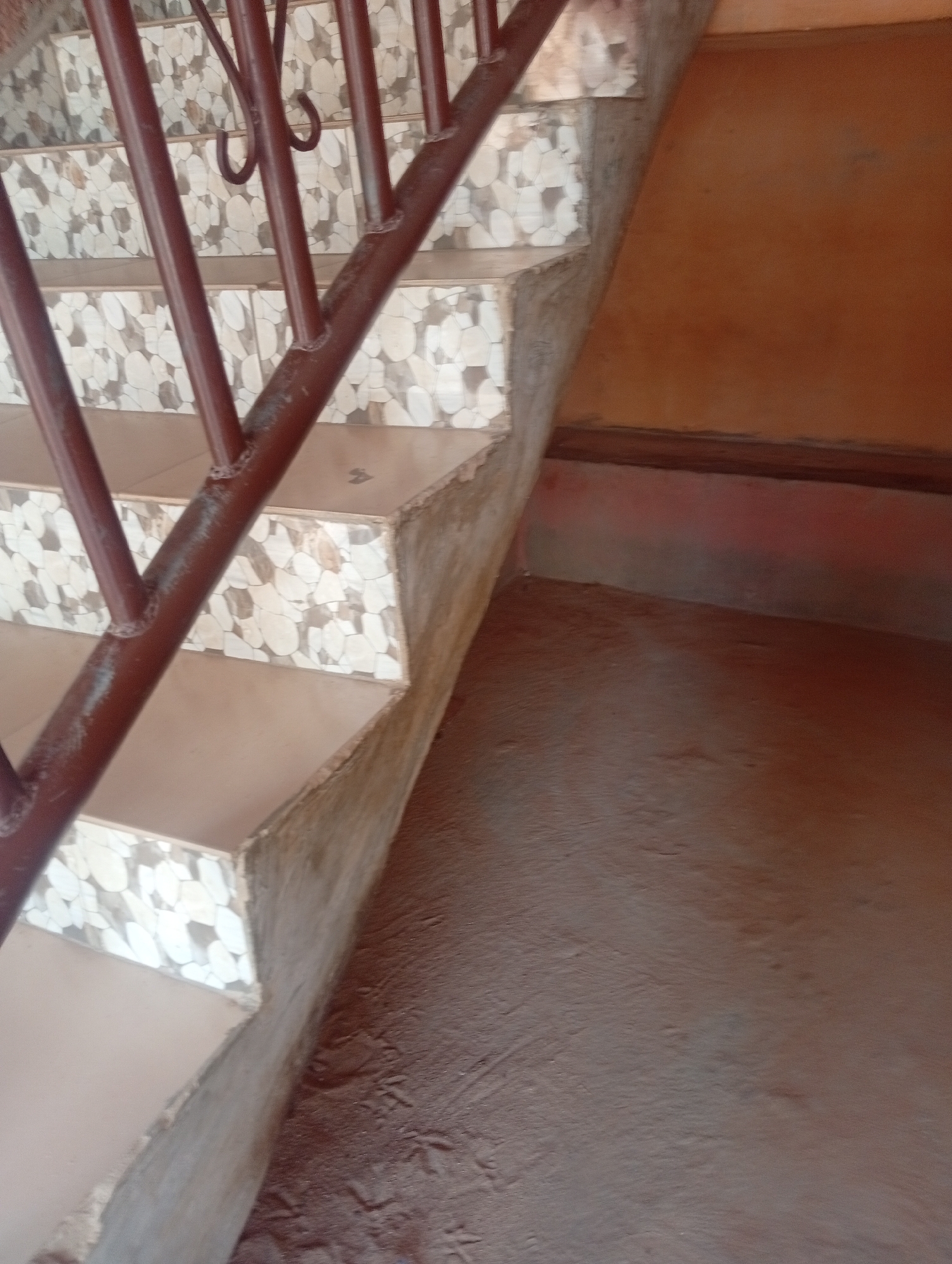 The image shows a close-up view of a set of stairs with a brown metal railing. The stairs have light-colored tiles with a patterned design on the risers, and the treads appear to be a neutral color. The sides of the stairs are made of concrete. To the right of the stairs, there is a wall painted a warm, orange-brown color. The floor in front of the stairs is a reddish-brown, textured surface. The lighting suggests it is daytime, and the overall setting appears to be outdoors or in a semi-outdoor area. There are no people visible in the image, and no text is present. The scene depicts a functional part of a building, likely a staircase, with simple and somewhat rustic finishes. The image is cropped to focus on the details of the stairs and the adjacent wall and floor.