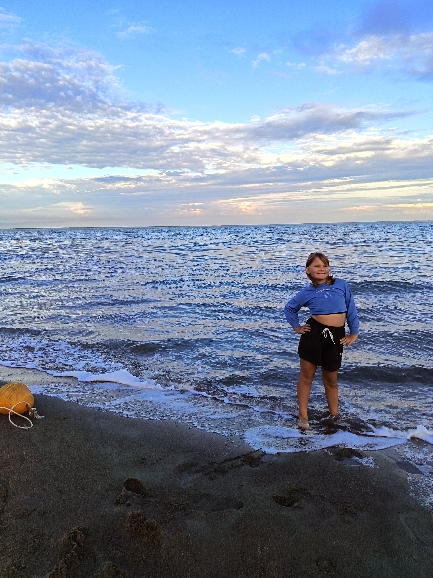A young girl stands on a dark sandy beach at the edge of a calm, blue sea, with waves lapping around her ankles. She is wearing a light blue, long-sleeved crop top and black shorts. Her hands are on her hips, and she is looking off to the side with a slight smile, appearing relaxed and content.
The sky above is a vibrant blue, with scattered white and gray clouds, suggesting it is either early morning or late afternoon. The water is a deep blue, reflecting the sky, and small white foam trails mark the movement of the waves. In the foreground, to the left of the girl, a bright orange buoy is partially visible on the sand, tethered by a white rope. The sand itself is dark and appears wet near the water's edge, with footprints visible further up the beach.
The scene conveys a peaceful and serene atmosphere. The lighting suggests a calm day, possibly during golden hour. The location cues point to a seaside setting, likely a beach. There is no visible text in the image.