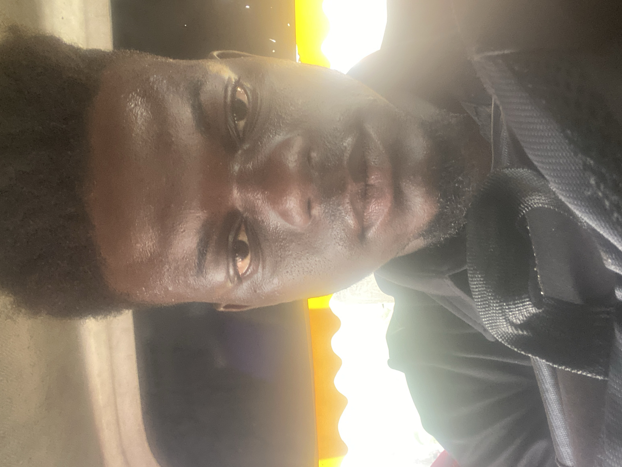 The image is a portrait of a dark-skinned man, rotated 90 degrees counter-clockwise, showing him looking directly at the camera. He has short, dark, kinky hair and a short beard. His face appears slightly shiny, possibly due to sweat or natural oils, particularly on his forehead and the bridge of his nose.
He is wearing a dark-colored garment, likely a black shirt or jacket. A black, woven fabric strap, possibly from a backpack or bag, is visible across his right shoulder (when the image is correctly oriented).
The background is composed of contrasting light and dark areas. A very bright, yellow-orange light source is prominent on the left side of the frame (bottom if rotated correctly), which creates a strong glow and causes some areas to be overexposed. This bright section has a distinctive wavy or scalloped edge. The right side of the background (top if rotated correctly) is mostly dark.
The strong light suggests it is daytime. The man has a neutral to slightly serious expression. He appears to be posing for a photograph or taking a selfie. There is no visible text in the image.