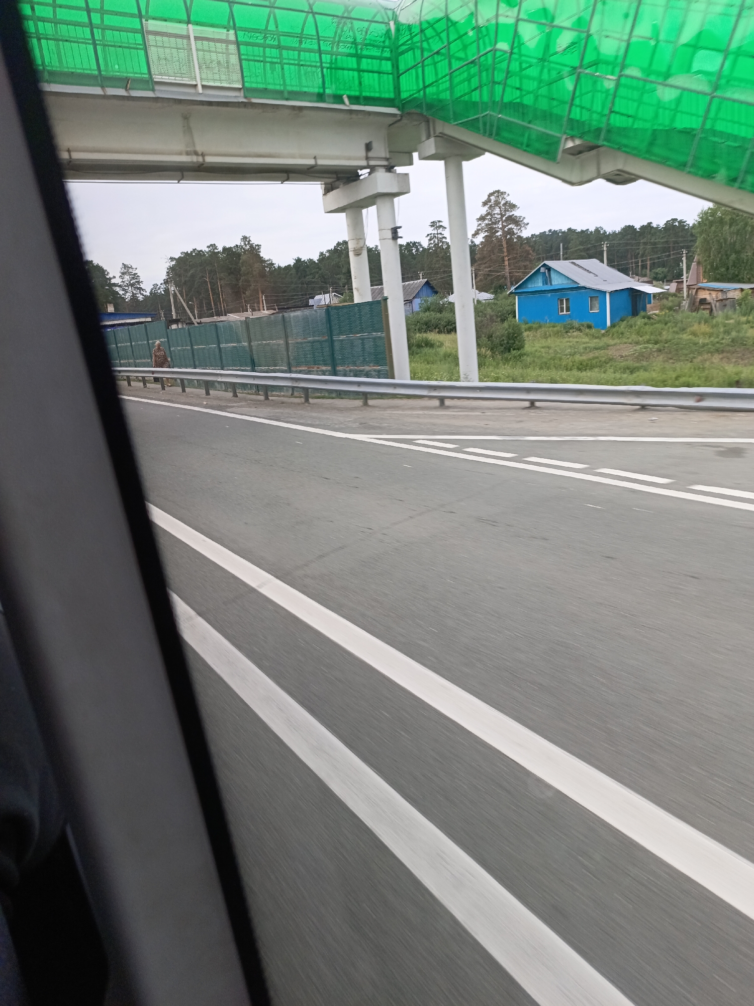 A view from a vehicle traveling on a road shows a green pedestrian overpass with a concrete structure supporting it overhead. The road surface is asphalt with white lane markings. To the right of the road, a turquoise metal fence runs along the side, backed by green grass and trees. A small blue house with a gray roof is visible amongst the greenery. Further in the distance, more trees form a treeline. The sky is overcast.