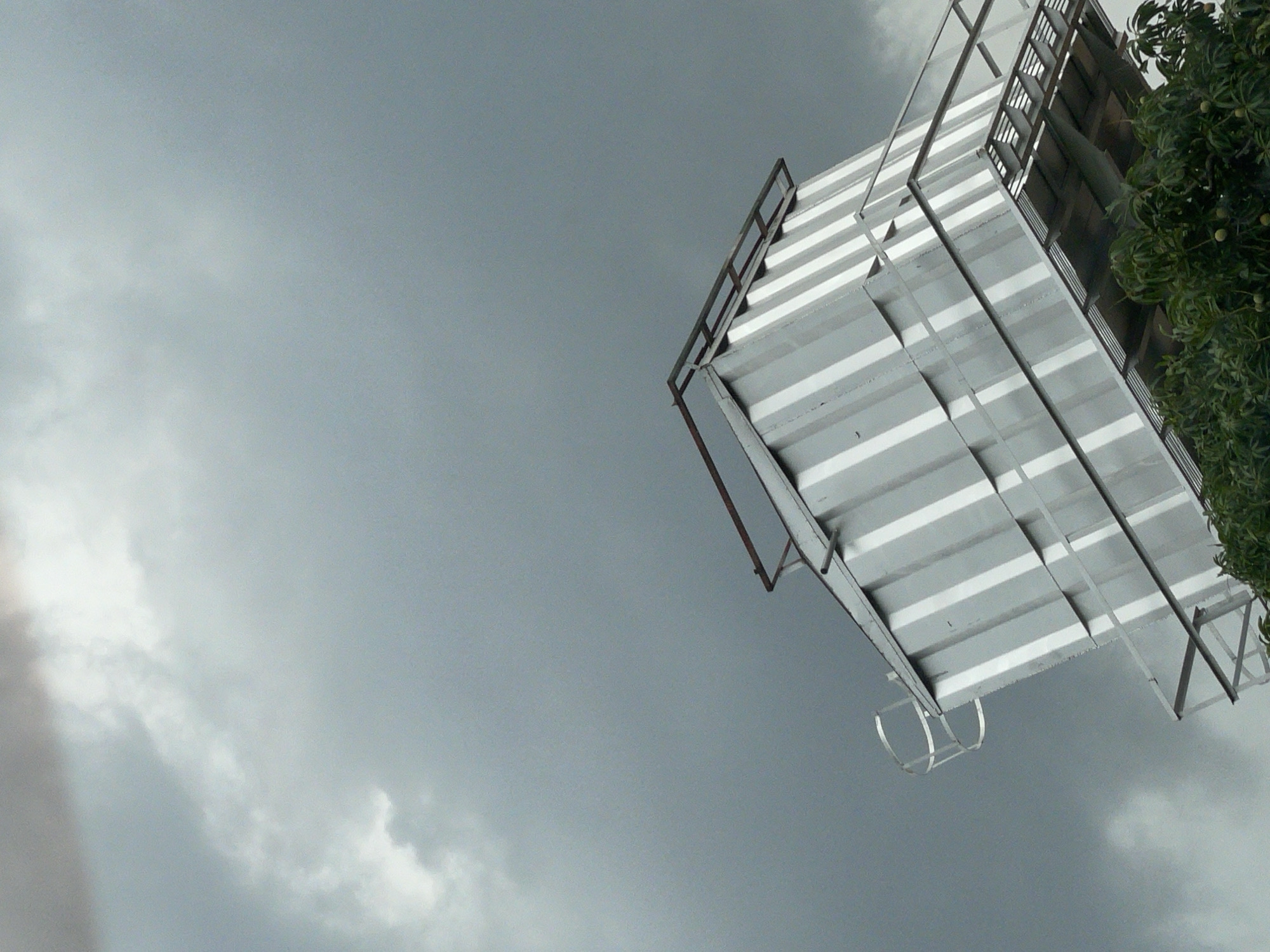 The image, likely taken in Jos, Nigeria based on the prompt, shows a low-angle view of the sky and a partially visible structure. The sky is overcast with grey clouds. The structure is rectangular, white and grey, with a metal frame, and appears to be an elevated platform or small building. Green foliage, possibly a tree, is visible to the right of the structure.