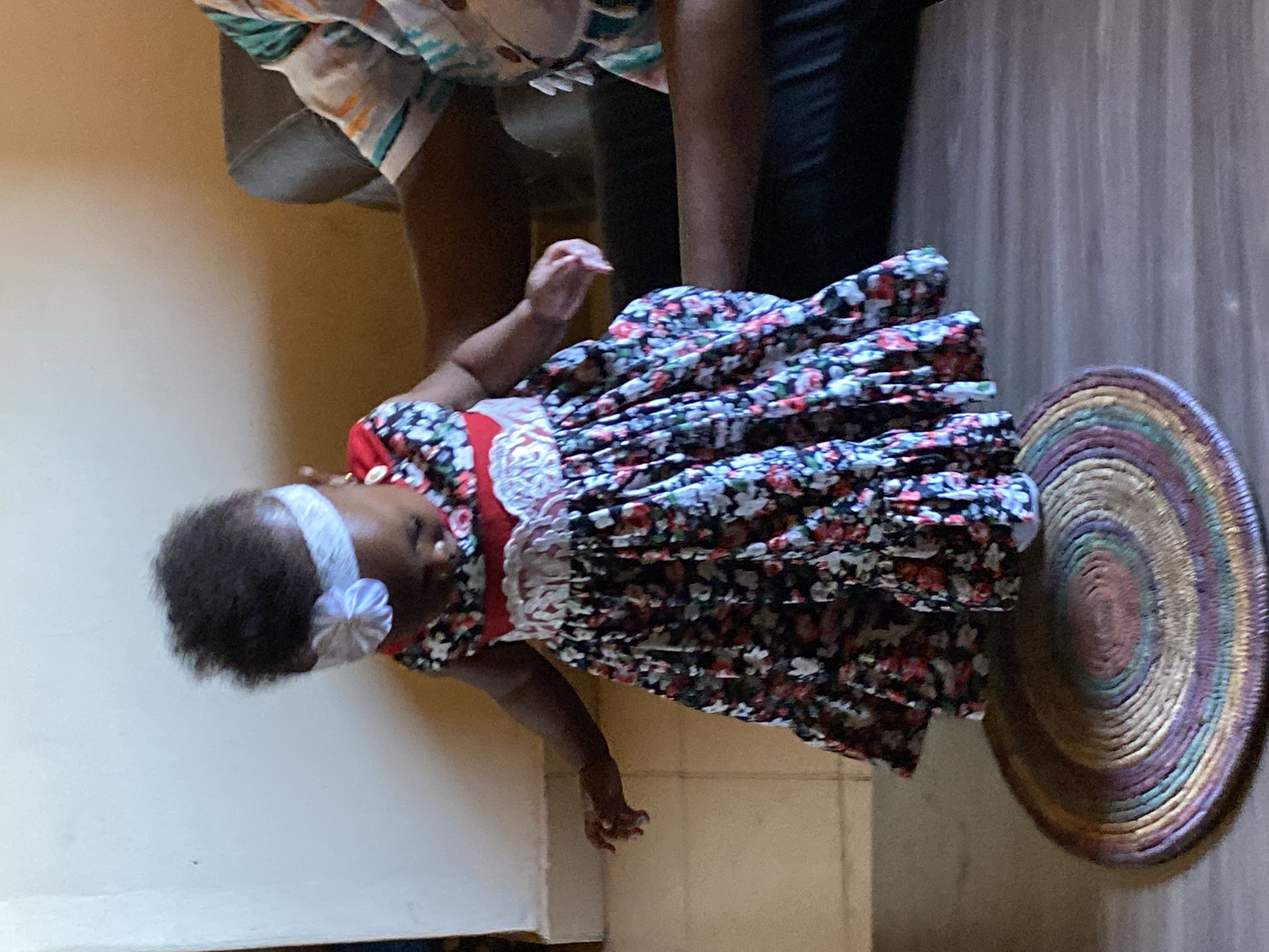 A baby with a white headband and a floral dress stands next to an adult on a wooden floor near a woven circular mat. The location is Bukuru, Nigeria.