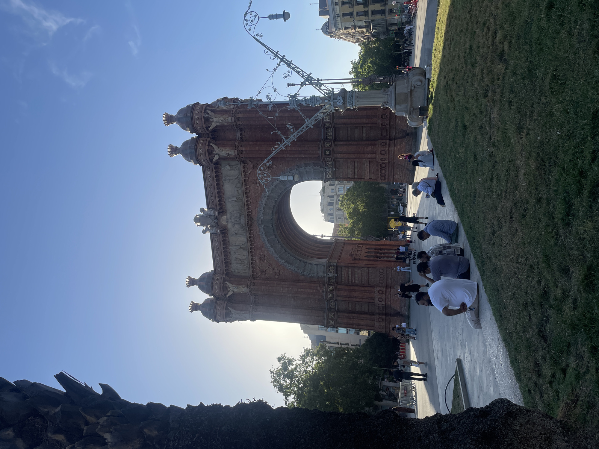 This media file captures the iconic Arc de Triomf in Barcelona, Spain, under a bright, clear blue sky. The grand brick archway, adorned with intricate sculptures and architectural details, stands prominently in the center of the frame. The sun, positioned low in the sky behind the arch, creates a strong backlighting effect, silhouetting parts of the structure and casting a brilliant glow, particularly through the main arch.
In the foreground and extending along the right side, a group of people is visible. Several individuals are sitting or kneeling on a paved pathway bordering a neatly trimmed grassy area, seemingly resting or observing the monument. One man in a white t-shirt and light-colored trousers is kneeling with his back partially to the viewer. Further back, additional people are seen walking and standing, alongside other urban elements like buildings, a decorative street lamp with elaborate metalwork, and distant trees.
The time of day appears to be late afternoon or early morning, indicated by the low sun angle and the bright, yet soft, quality of light. The weather is sunny and clear, with only a few faint clouds visible in the vast blue sky. The overall atmosphere is calm and relaxed, with visitors enjoying the public space around the landmark. In the bottom left corner, dark, textured foliage, possibly from a palm tree, partially frames the scene. No discernible text is visible on the arch or surrounding elements.