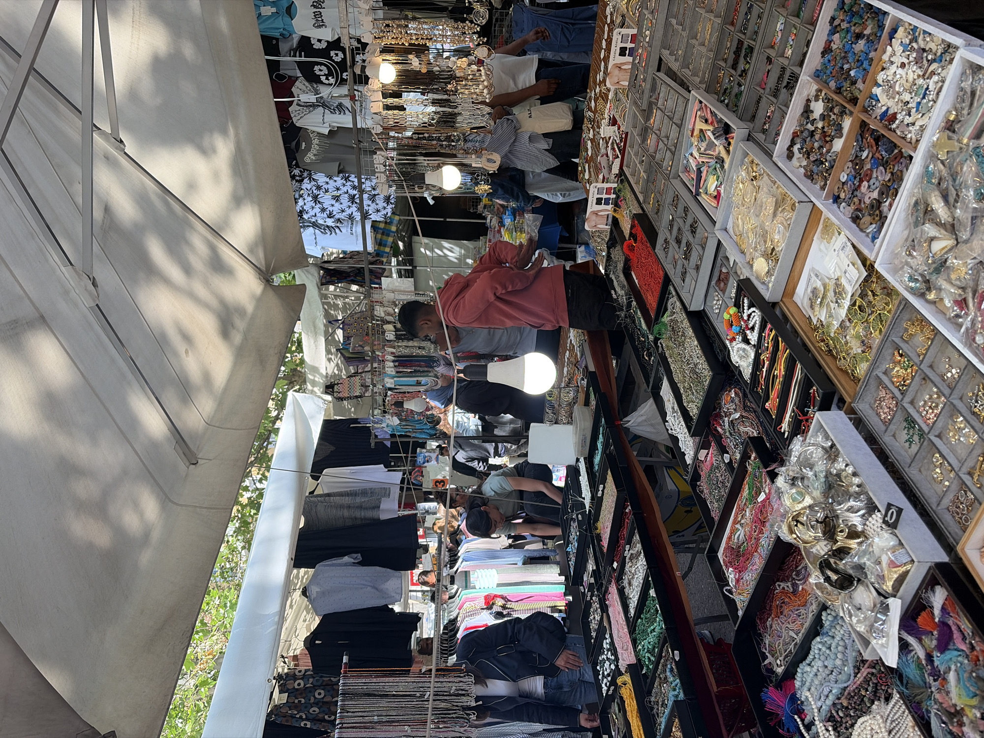 The image shows a street market scene, presumably in Milan, Italy. The market is under a white canopy that shades the wares and customers. Two rows of vendors have set up stalls. The stall on the left appears to be selling clothing, with shirts and pants visible. The stall on the right features various jewelry items, displayed in trays and boxes. Several people are browsing and shopping. One man in a pink hooded sweater and dark pants is actively examining items at the jewelry stall. Other individuals are seen looking at the clothes and interacting with each other. The environment seems to be a sunny day, with the white canopy providing shade and the merchandise well-lit.