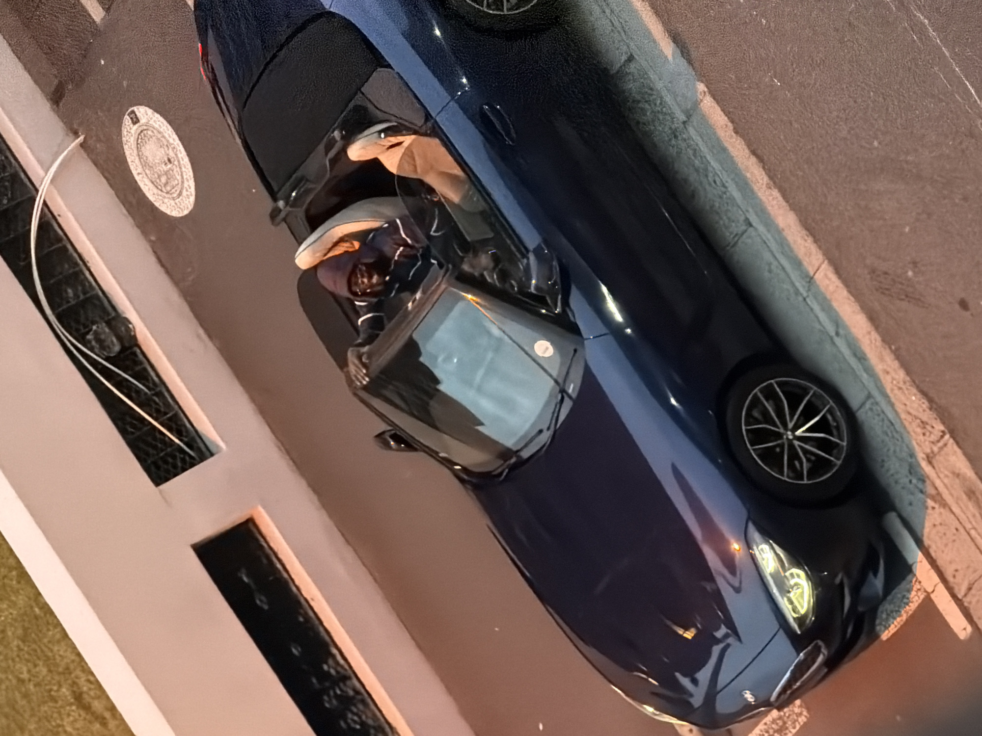 The image shows a dark blue convertible car parked on the side of a street next to a building. A person is sitting in the driver's seat. The building has distinctive decorative window grilles and a round, patterned plaque. The scene is likely in the city of Cape Town, South Africa.
