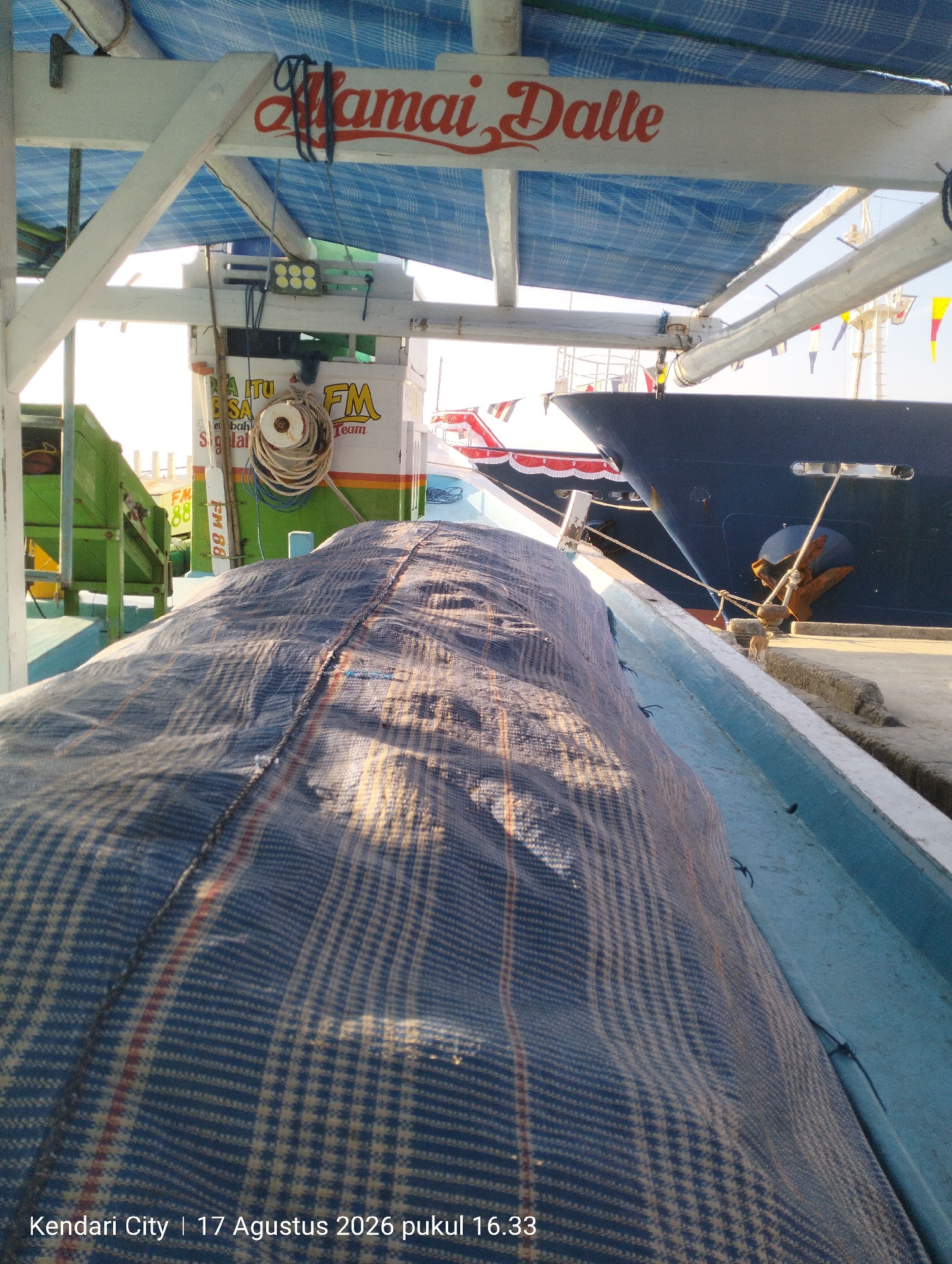 The image depicts a close-up view of a fishing boat docked in Kendari City, Indonesia, on August 17, 2026, at 16:33. The boat is covered with a blue and tan checkered tarp, suggesting cargo or equipment is being protected. In the background, a larger, dark blue ship is visible, with its anchor deployed. The boat appears to be on a concrete dock, with some wooden structures and a blue tarp overhead. The text on the boat indicates it is named "Mamai Dalle" and features "FM Team" branding. The scene is brightly lit, suggesting it is daytime.