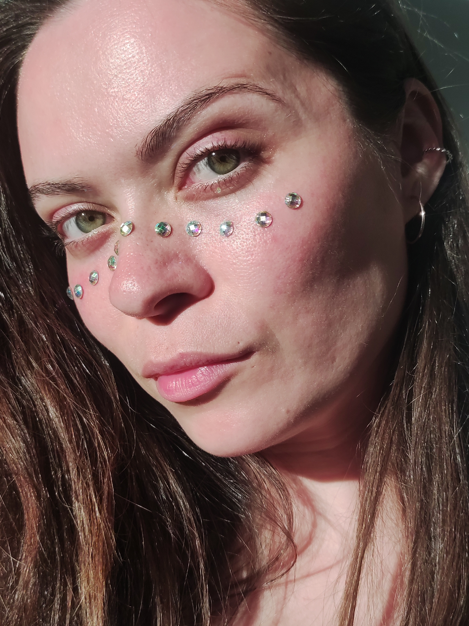 A close-up photograph shows a person with fair skin and green eyes, adorned with small, iridescent rhinestones on their face. The rhinestones are arranged along the cheekbone, nose, and forehead. The person has long brown hair. The background is indistinct, with variations in light suggesting an indoor setting with possible natural light sources. The specific location is not discernible from the provided information.