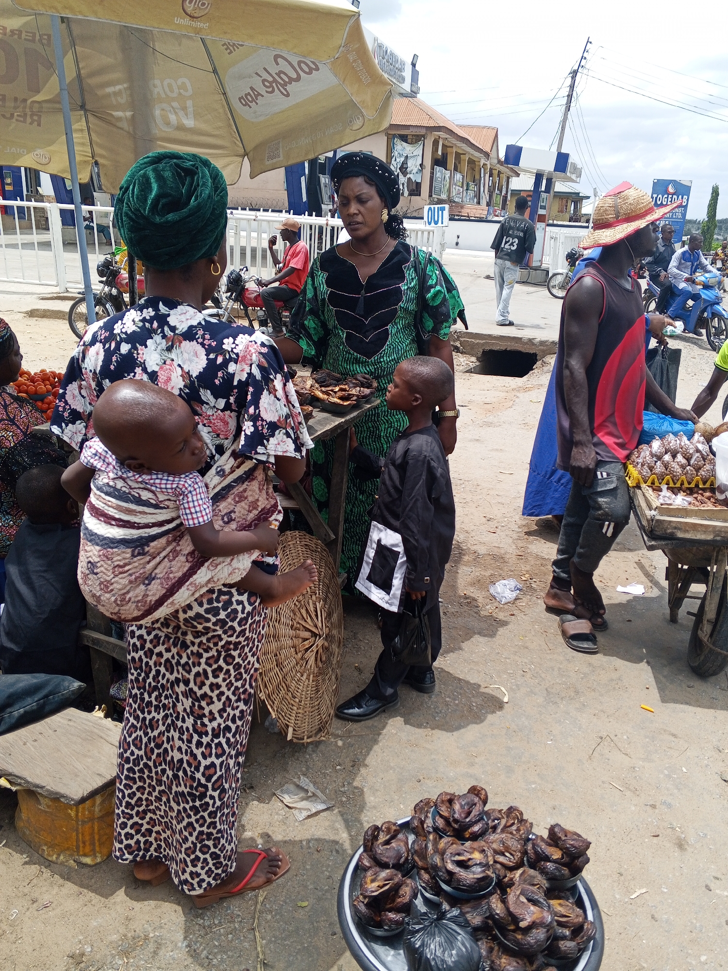The image captures a street scene in Zango-ndaji, Nigeria. In the foreground, a woman with a baby secured on her back in a striped and patterned wrap, is wearing a floral top and leopard-print skirt. Nearby, a woman in a green patterned dress interacts with a young boy in a black suit. To the right, a man wearing a straw hat and a tank top pushes a cart filled with eggs. A metal tray filled with what appears to be smoked fish sits on the ground in the lower foreground. People and motorcycles are visible in the background, along with buildings and signage, including an advertisement. The area seems to be a market or roadside vendor setting.