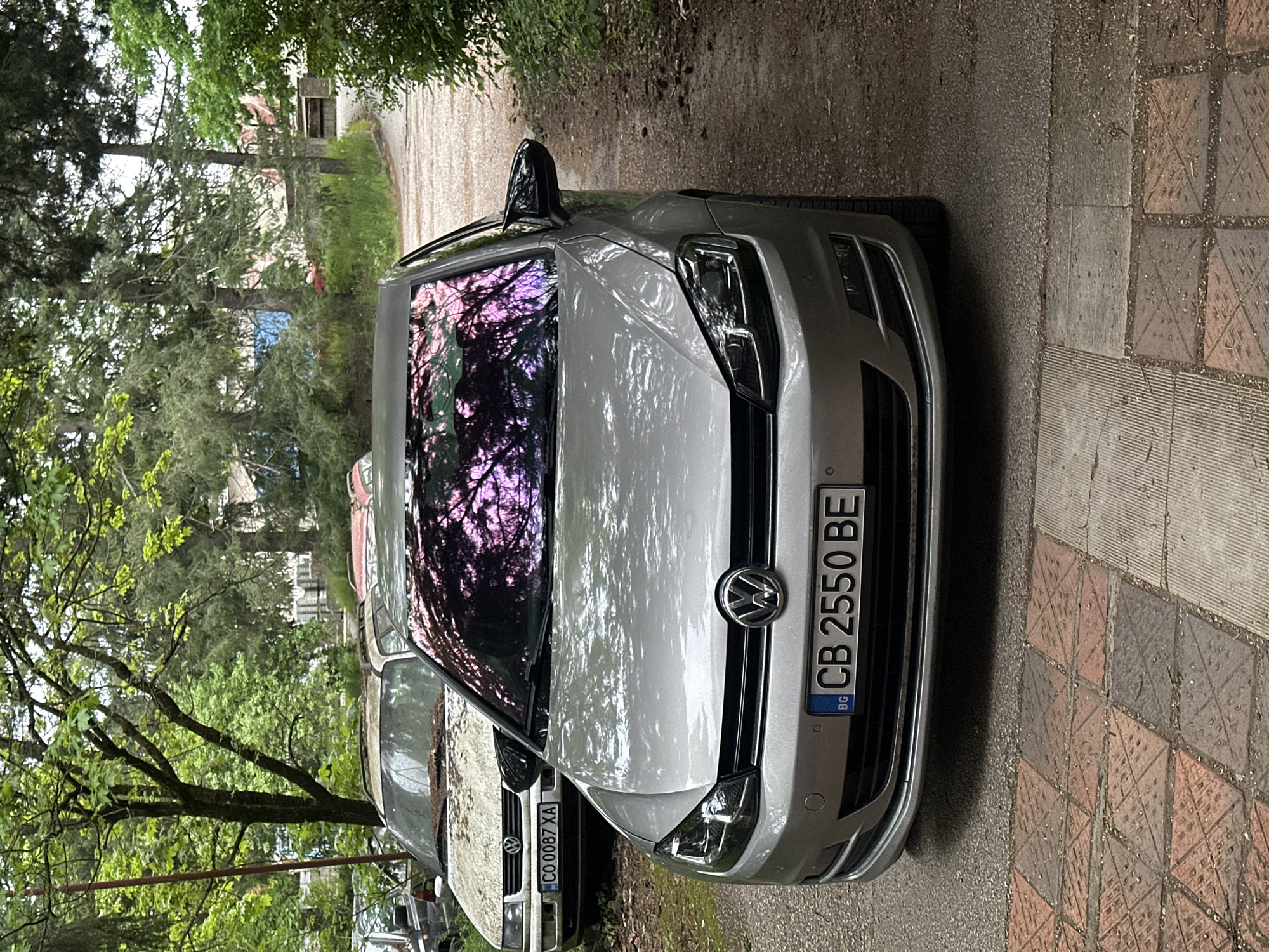 The image shows the front of a silver Volkswagen Golf parked on a narrow road with a brick-patterned sidewalk to the right. The car's license plate reads "CB 2550 BE". Another older, dirtier Volkswagen vehicle is partially visible behind the Golf, with its license plate reading "CO 0087 XA". The windshield of the silver Golf reflects the surrounding trees. The location appears to be a residential area, with trees and greenery visible in the background.